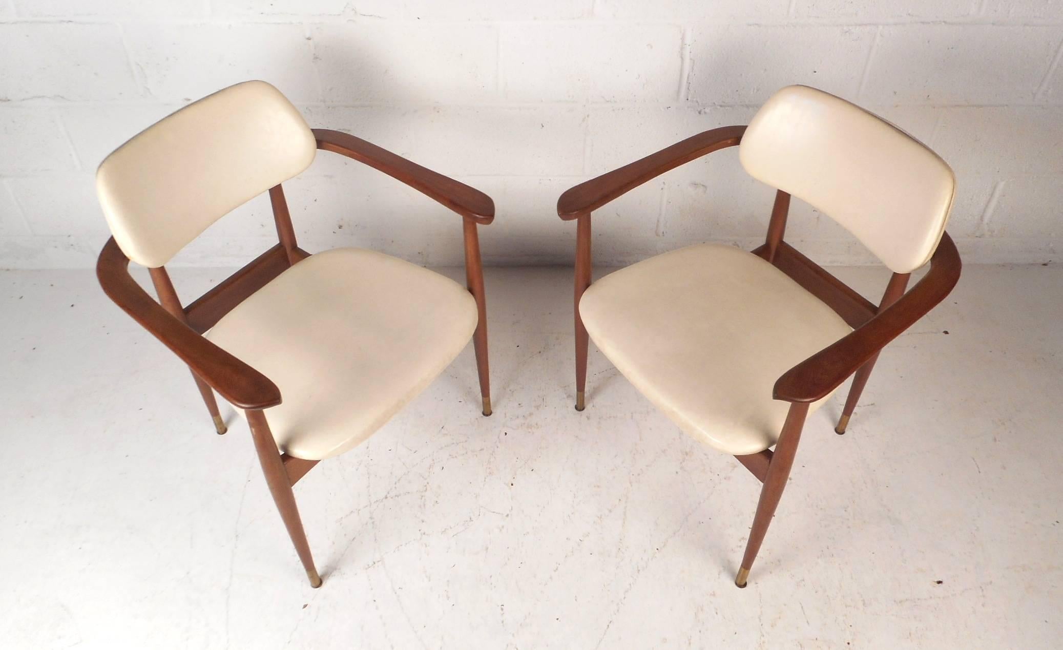 This stunning pair of vintage modern chairs feature sculpted arm rests, a floating style backrest, and tapered legs. The sleek and comfortable design has thick padded seats covered in white vinyl. This attractive pair of side chairs have brass