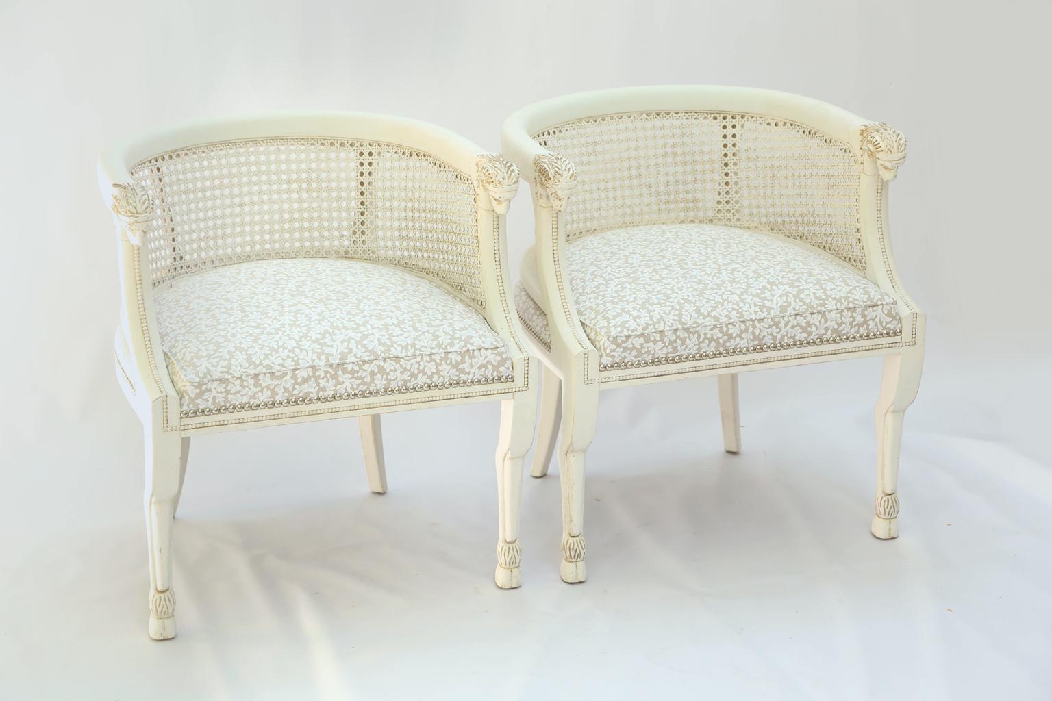 Pair of barrel-form, painted bergère chairs, each having a rounded, molded crestrail, extending into carved ram's masks, over hand-caned backs and sides, crown seat upholstered in vintage floral-printed linen with silver nailheads, raised on front