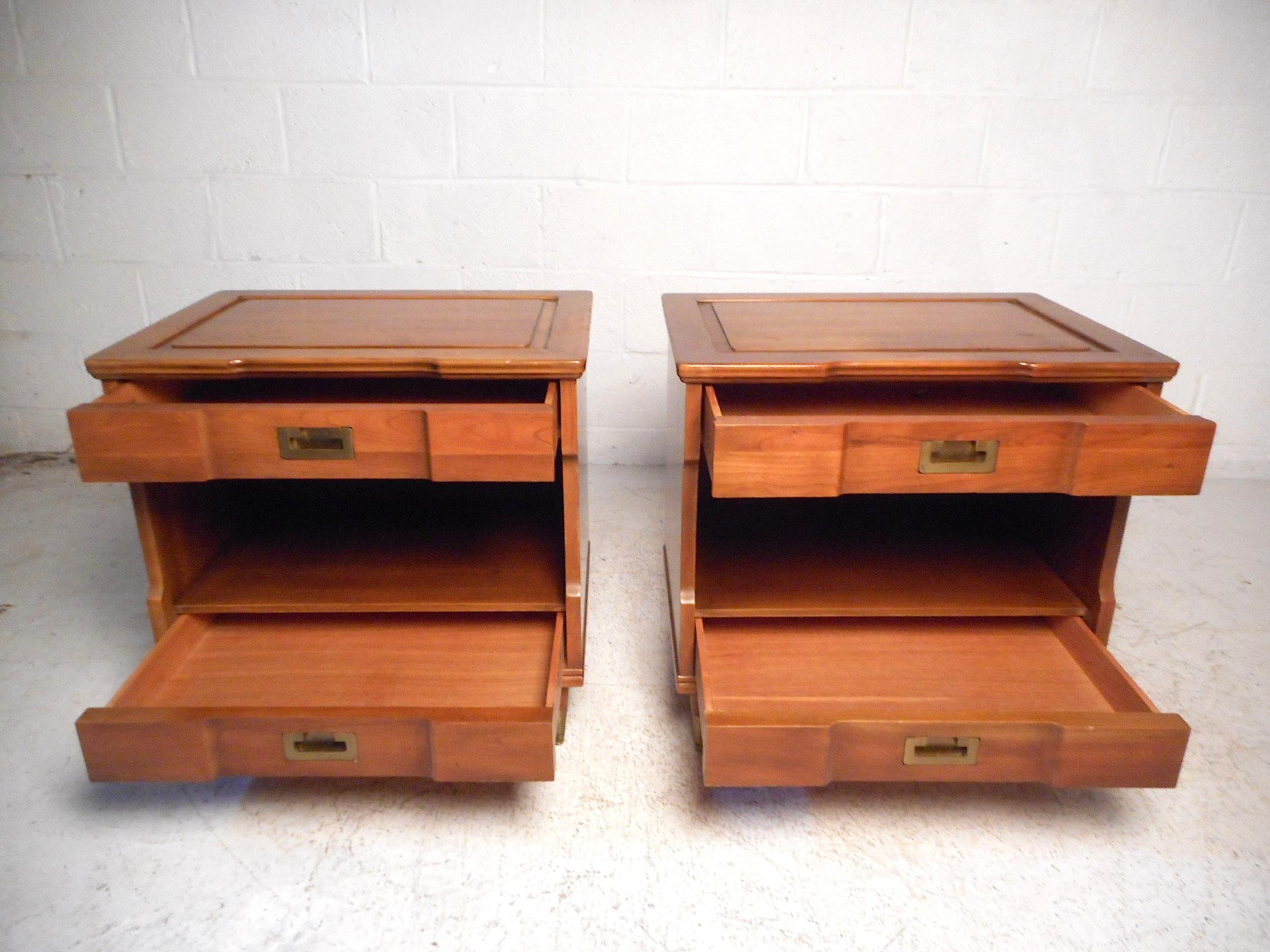This stylish pair of vintage modern nightstands feature a sturdy walnut construction with a lightly colored finish, two drawers with dovetail wood-joints signifying quality craftsmanship while offering ample storage space, brass Campaign style