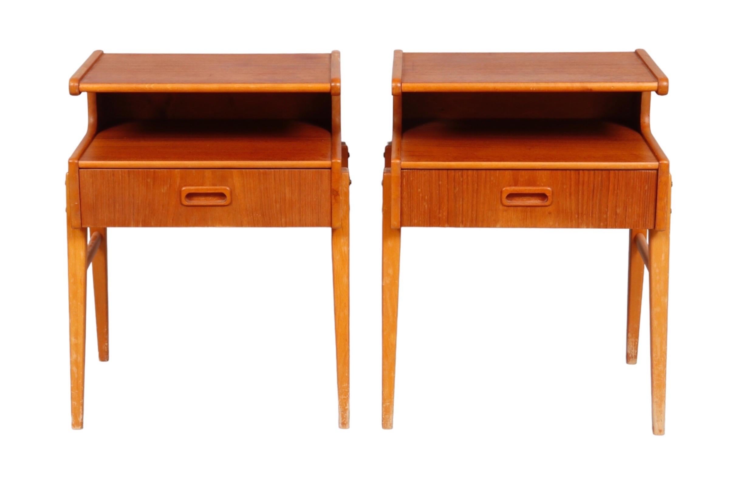A pair of mid century nightstands. Frames are made of beech with teak veneer. Each has a small shelf above a single drawer that opens with a recessed handle.