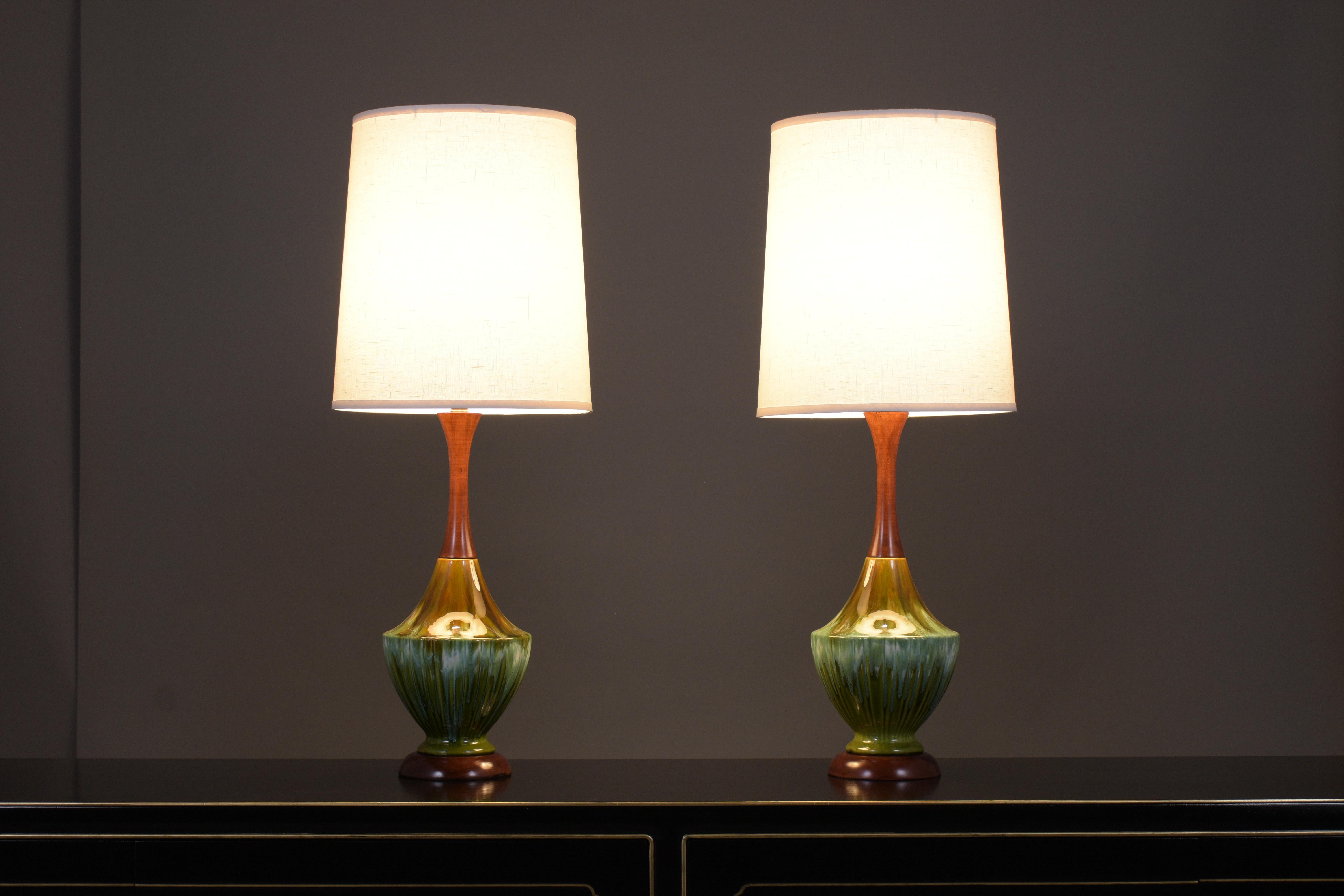 An extraordinary pair of 1960s mid-century table lamps that feature a porcelain vase painted in a green and amber color combination with a beautiful glazed finish. The lamp sits on a carved circular base in mahogany color with a lacquer finish and