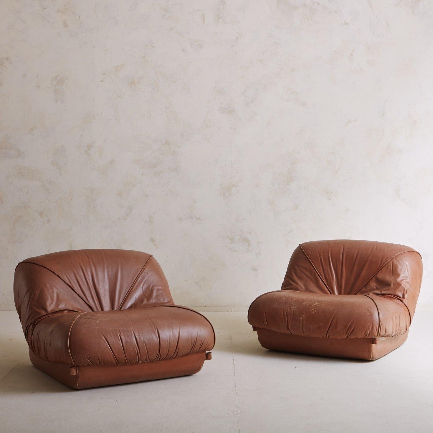 A pair of mid century lounge chairs featuring dramatically ruched seats and backs. These chairs retain their original cognac leather upholstery and the inset bases sit directly on the ground. Sourced in France, 1970s.