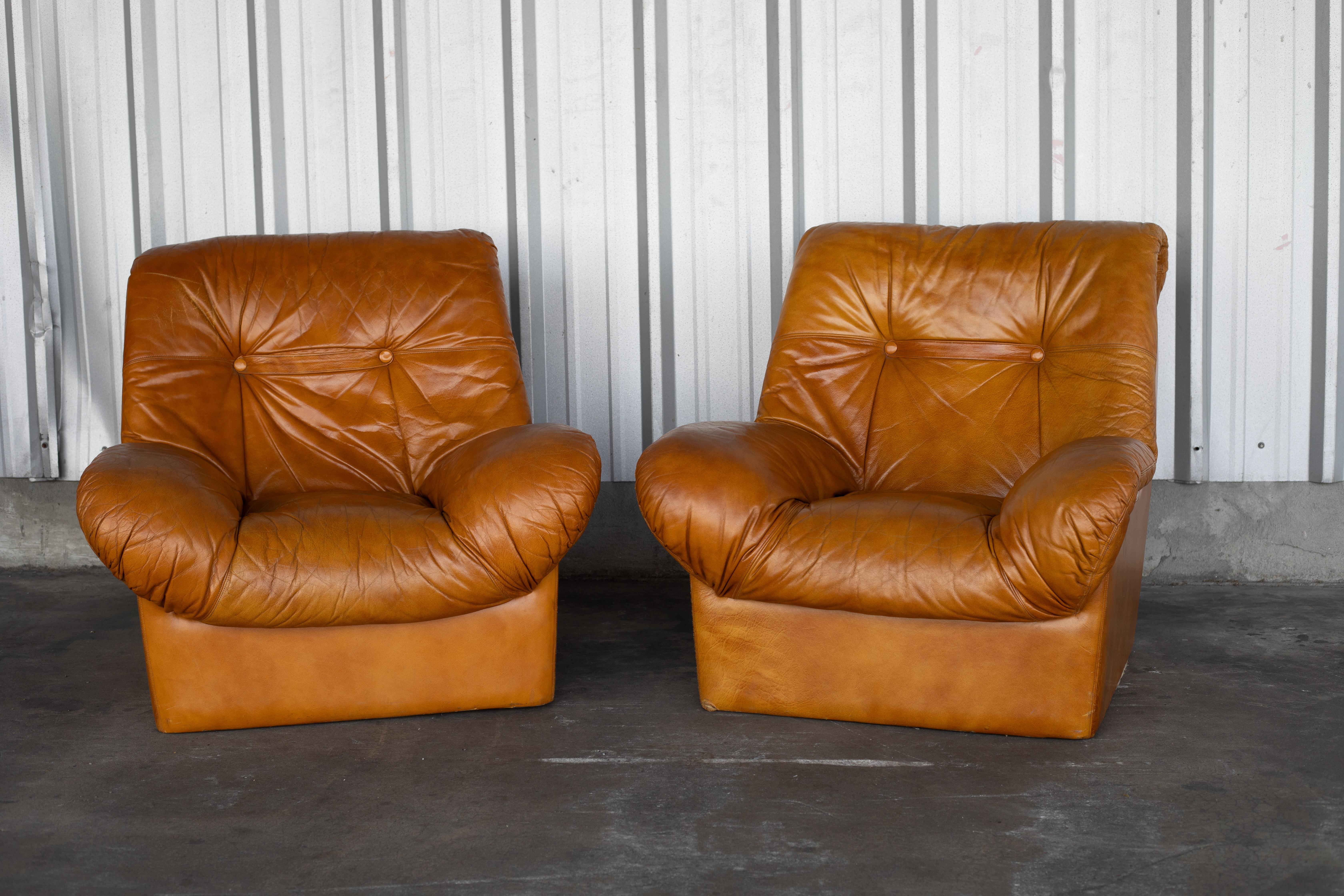 A pair of Mid-Century Scandinavian tan leather lounge chairs. These plush vintage lounge chairs feature deep cushioning, oversized arms, and a low, sculptural profile. Accented with tufted button details on the backrest, this 1970s design embodies