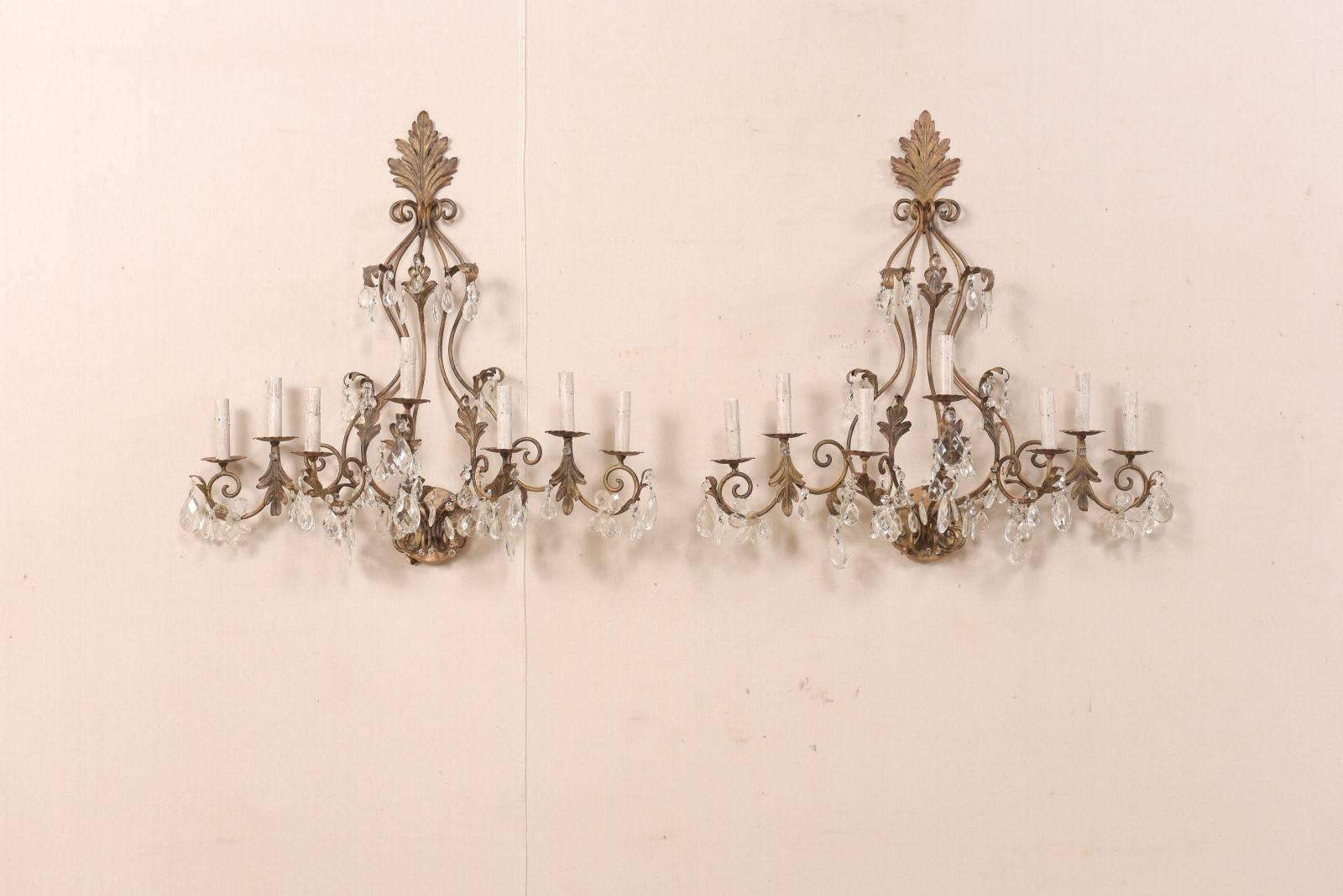 A pair of French crystal seven-light sconces from the mid-20th century. This pair of generously-sized, midcentury, French scones feature a warm bronze hue iron armature with scroll and leaf motif, which is adorn with various shapes of hanging