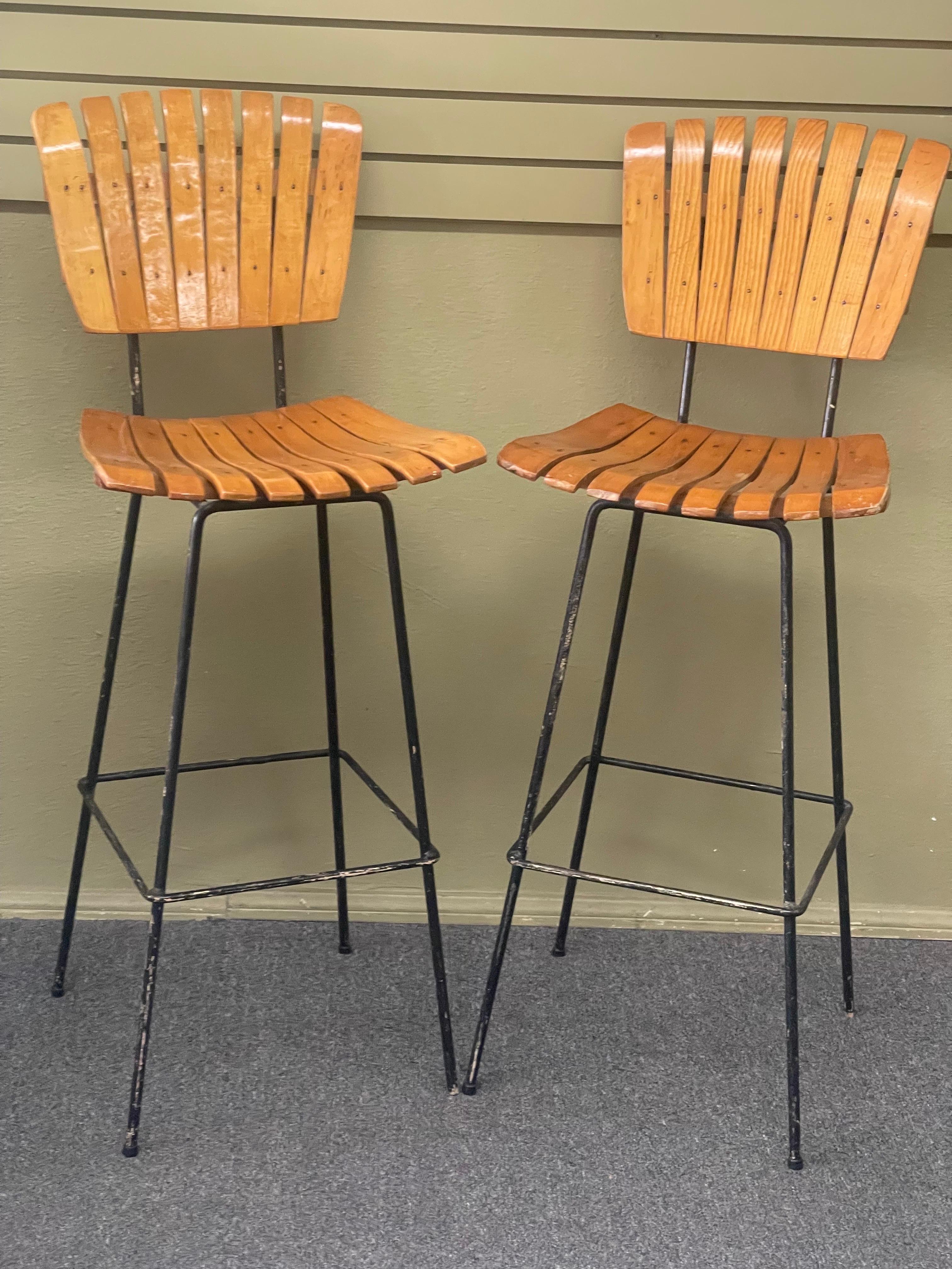 A cool pair of mid-century slat bar stools in the style of Arthur Umanoff, circa 1950s. The stools have a great look and are in well worn condition with a nice patina on the wood as well as the iron legs (please see pictures. Each bar stool measures