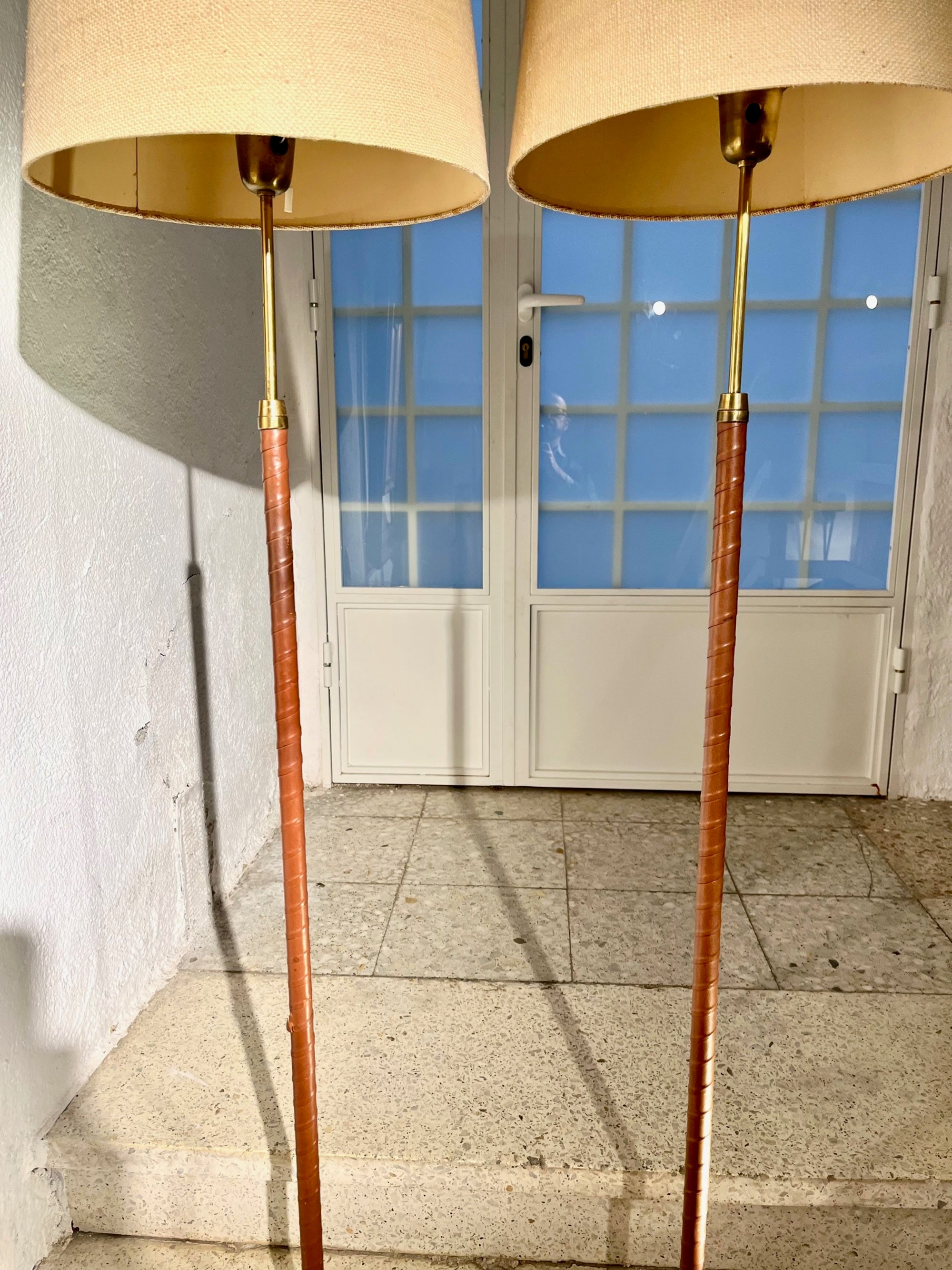 Elegant pair of mid-century Swedish floor lamps attributed to Einar Bäckström, circa 1950s.
Each lamp features a sleek, tapered column wrapped in fine cognac-colored leather, rising from a solid brass base with a warm, golden patina. The refined