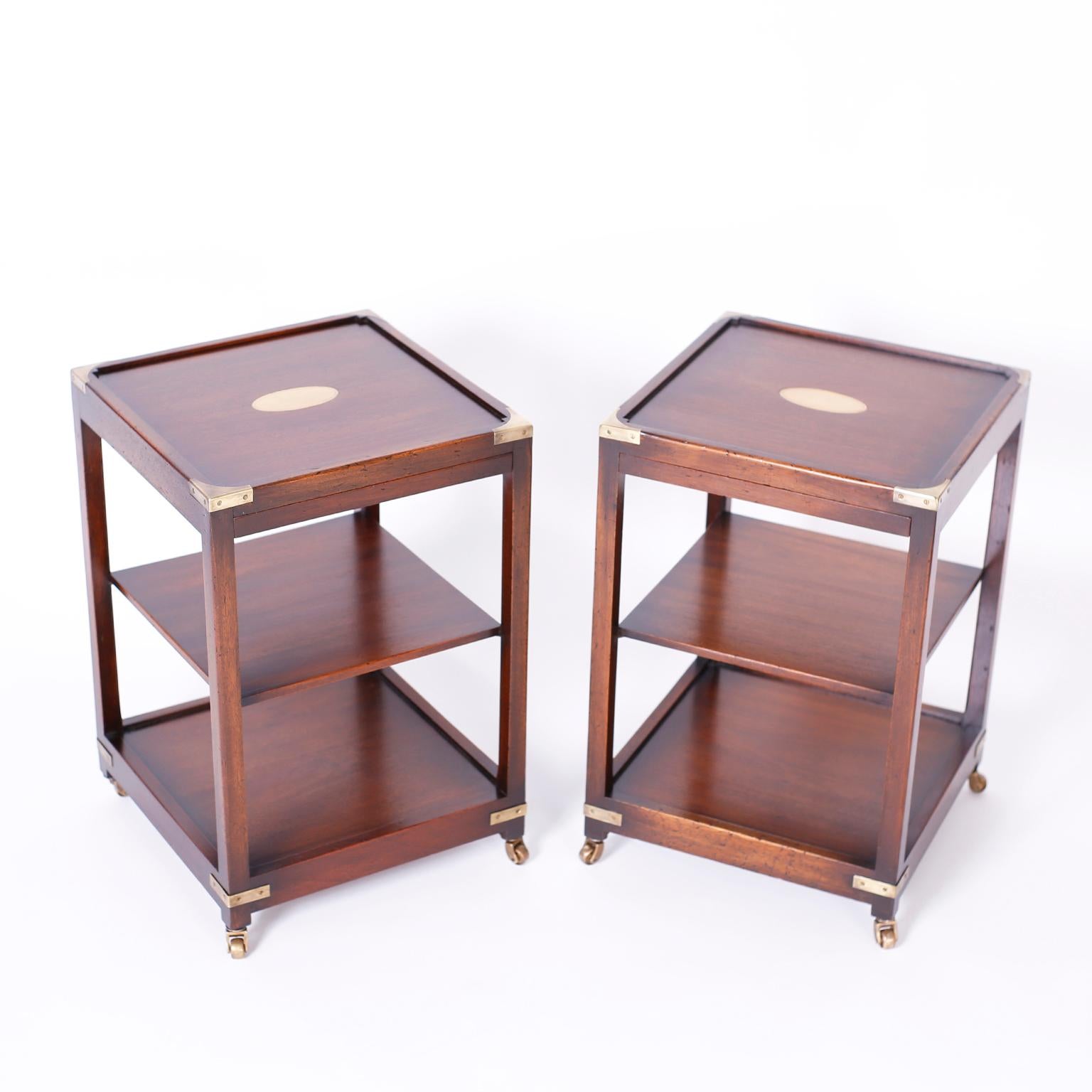 Handsome pair of midcentury Campaign side tables or stands crafted in mahogany with three tiers for plenty of storage, featuring brass hardware, casters and center oval plaque and pullout utility trays.