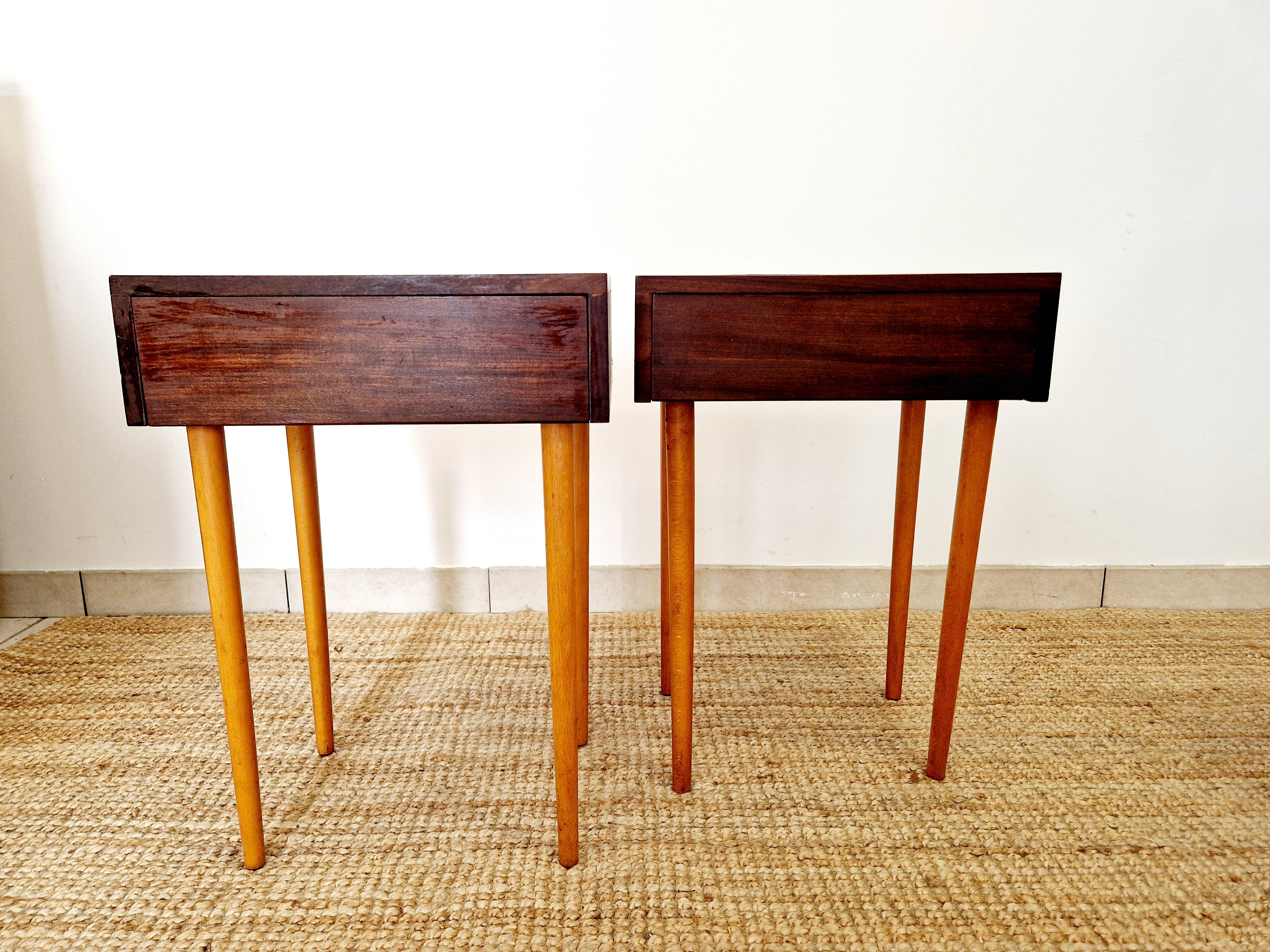 This pair of mid-century Tola bedside cabinets features a solid teak construction with a warm medium wood tone and teak handles, embodying the classic mid-century modern style popular in the 1960s. These fully assembled cabinets boast one drawer and