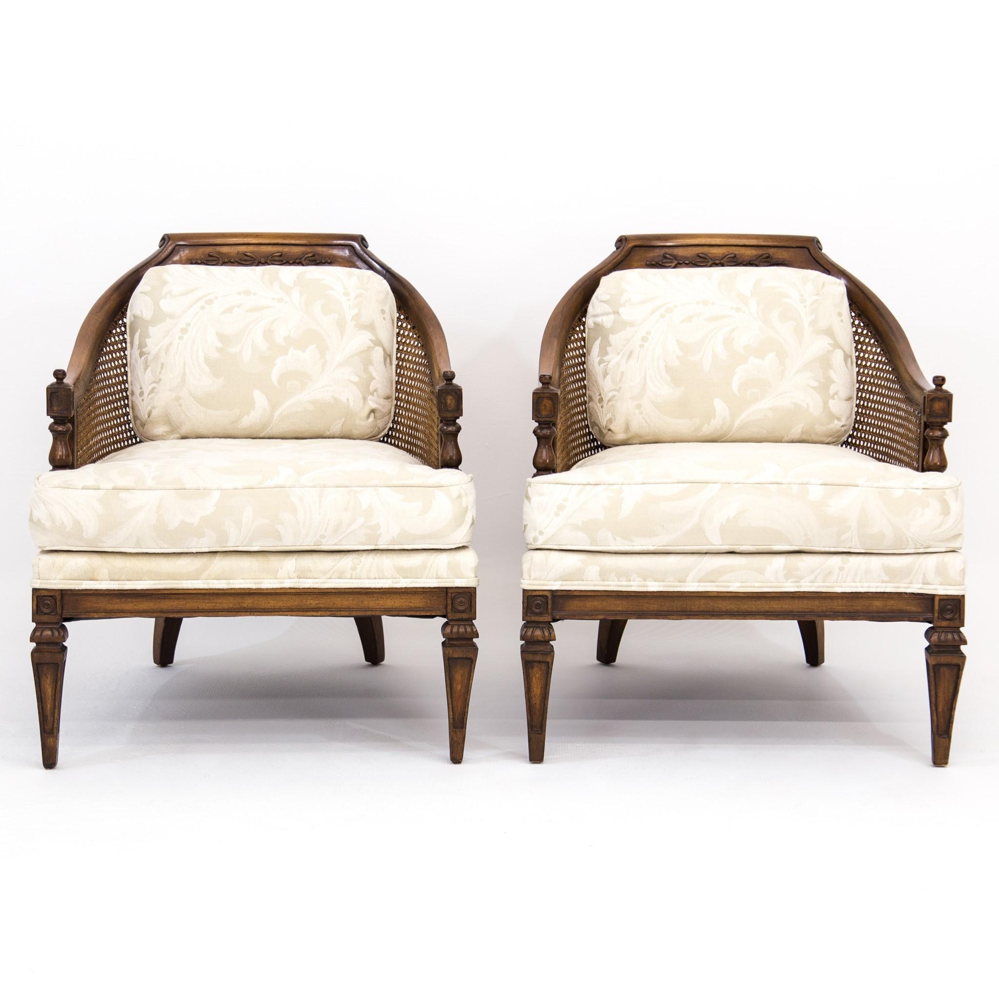Pair of walnut and caned barrel back tub lounge chairs with carved detail and ivory and cream brocade floral upholstery, circa 1970s. They feature carved and scrolled crested top rails ending in swoop arms in horseshoe form above Louis XVI-style