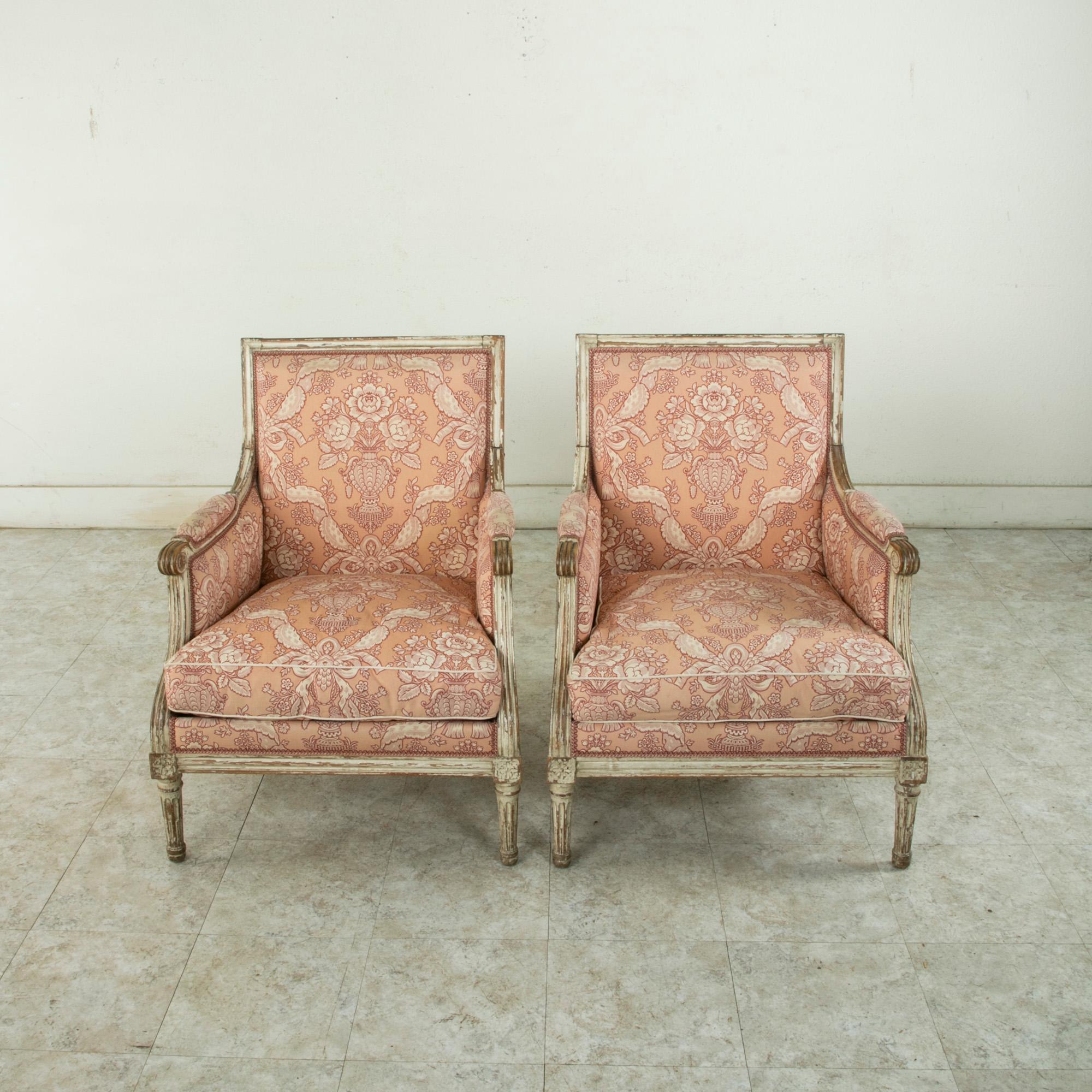 This pair of mid-twentieth century French Louis XVI style bergeres or armchairs features carved rosettes at the die joints and scrolling armrests. The chairs rest on tapered fluted legs. The seats, back, and arms are upholstered in a pink fabric