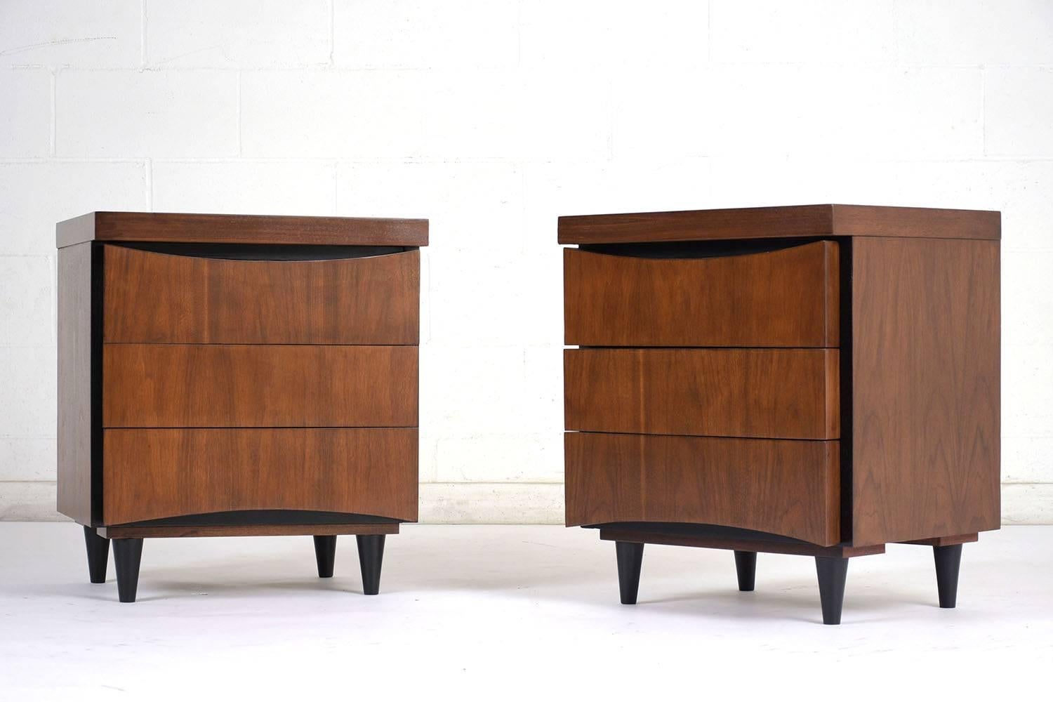 This pair of 1960s American of Martinsville nightstands features ample storage with three drawers. The nightstands have been completely restored and stained in a rich walnut color with black accents. The clean front of the nightstands is made