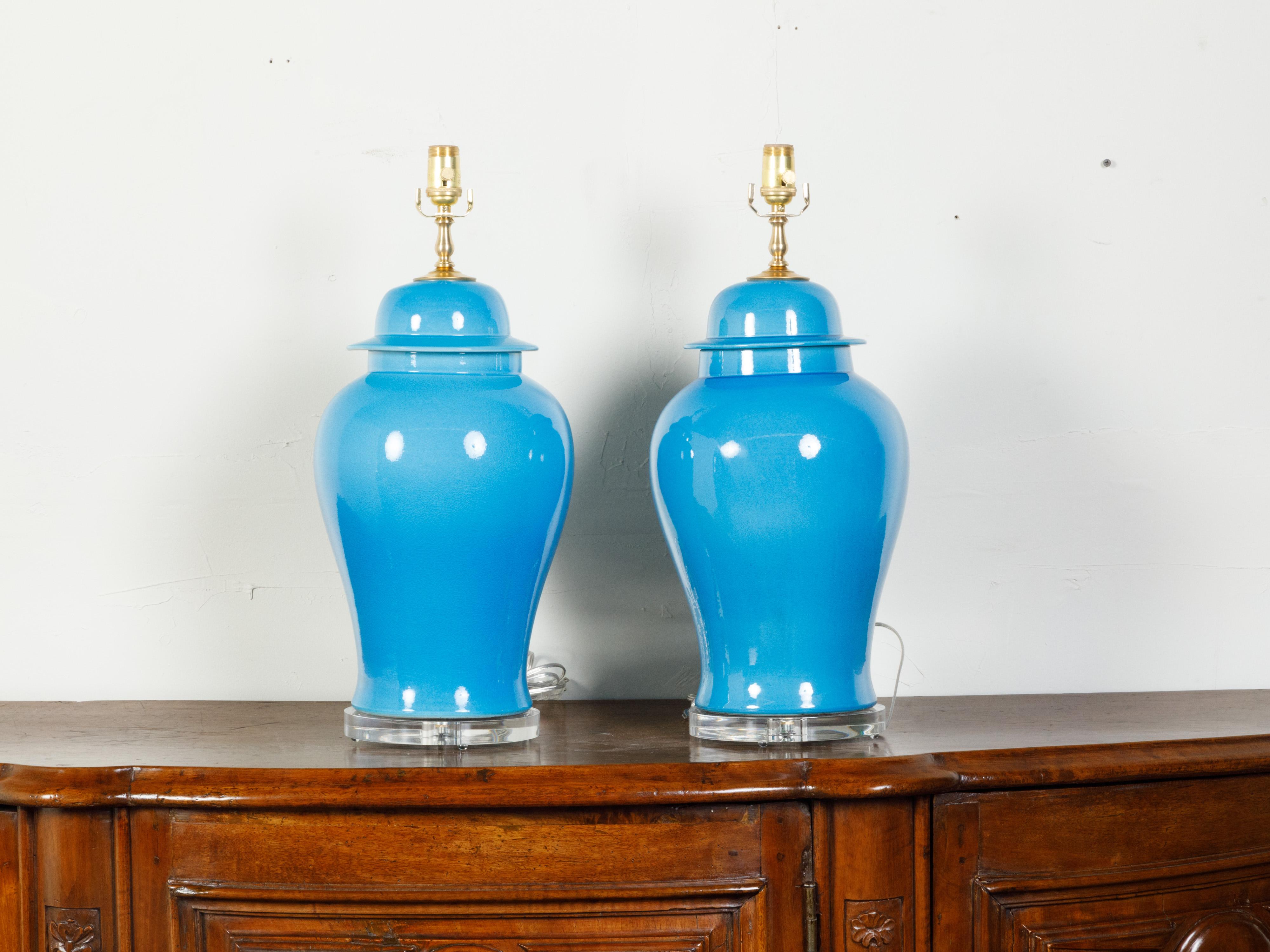 A pair of blue porcelain table lamps from the mid 20th century, made of vases mounted on lucite bases. Created during the midcentury period, each of this pair of table lamps features a blue porcelain vase topped with domed lids. Showcasing tapering