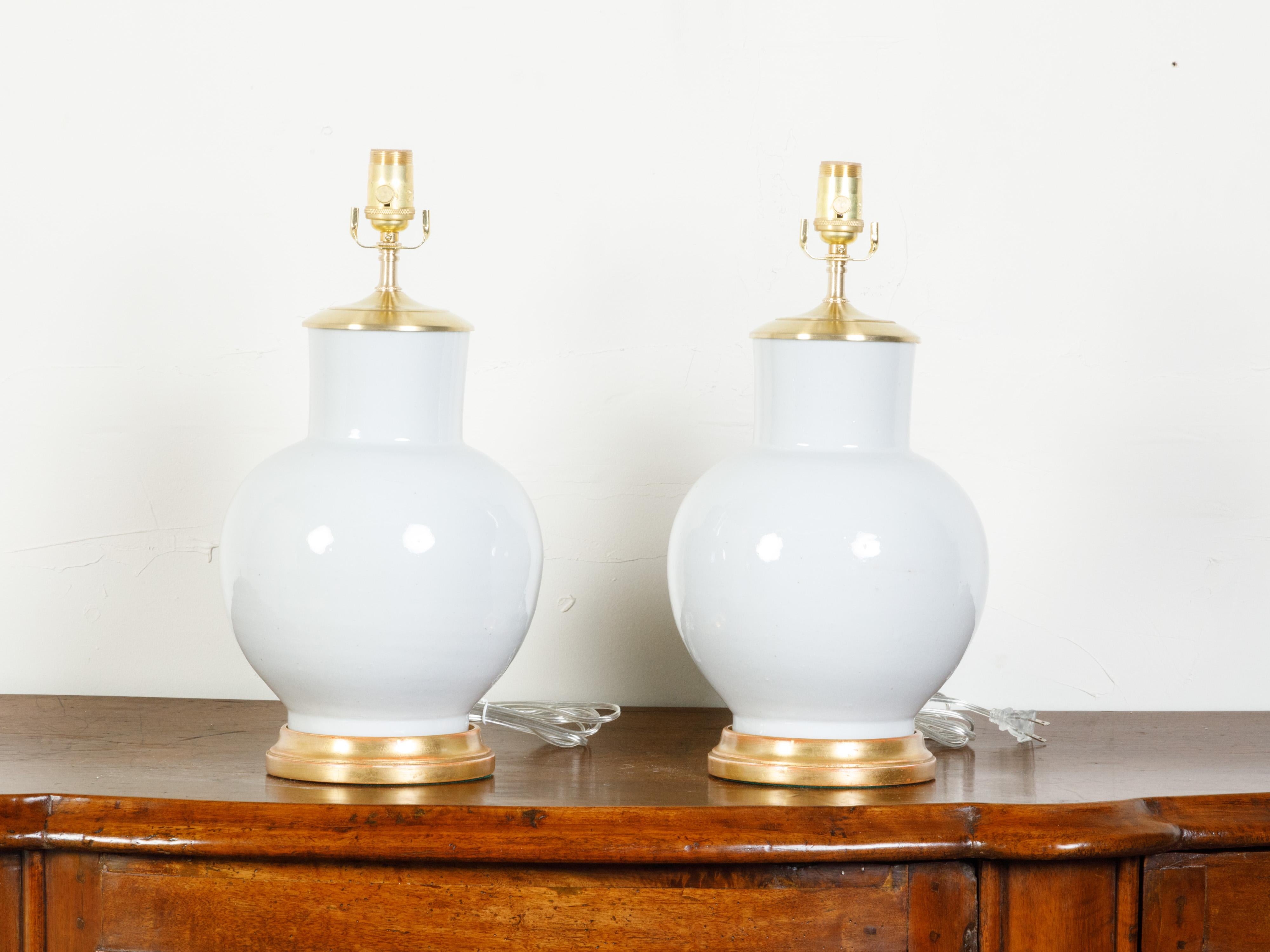 A pair of Chinese vintage white ceramic table lamps from the mid 20th century, with giltwood bases. Created in China during the Midcentury period, each of this pair of table lamps, professionally rewired for the US, features a generous white body