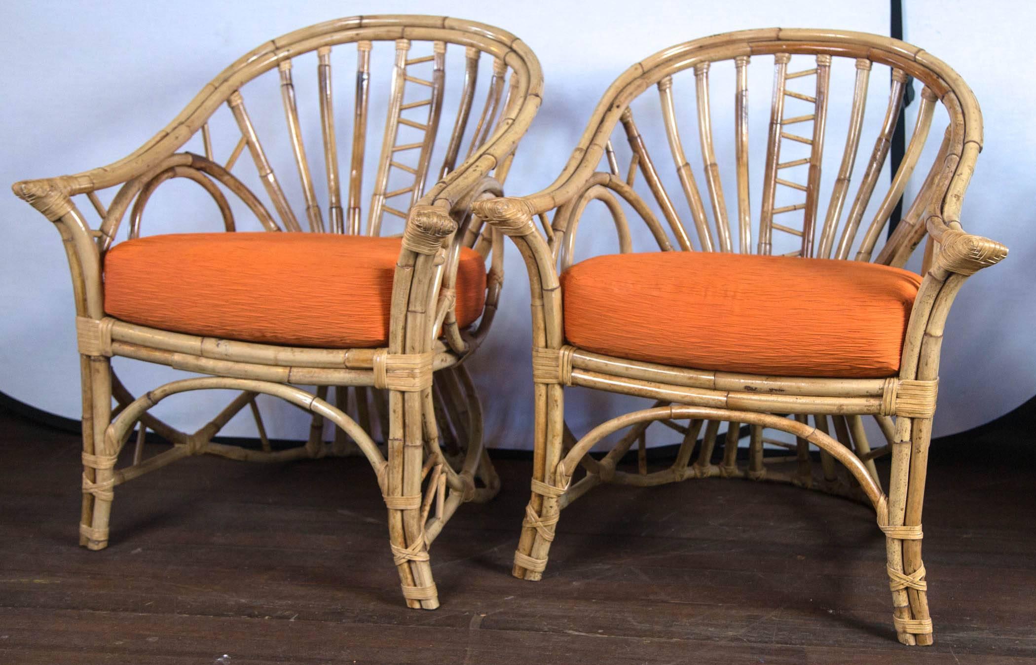 Pair of gracefully curved back rattan arm chairs with corset backs and circle sides. Two orange cushions.