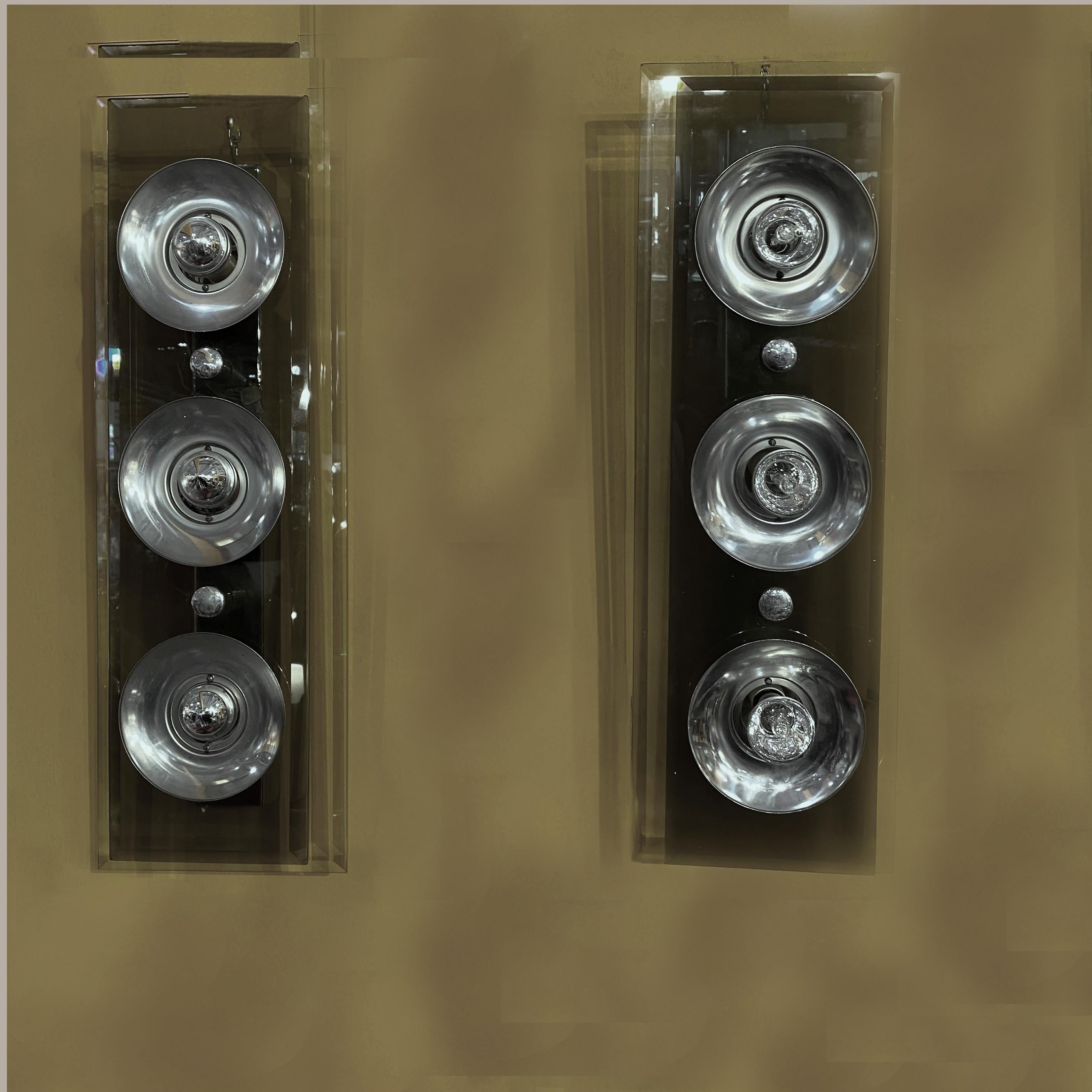 Paire d'appliques des années 1970 de forme rectangulaire imitant l'éclairage des projecteurs. Exécution élégante avec deux panneaux de verre dont un fumé. Les deux verres sont émaillés pour une apparence luxueuse.
Trois sources lumineuses sur chaque