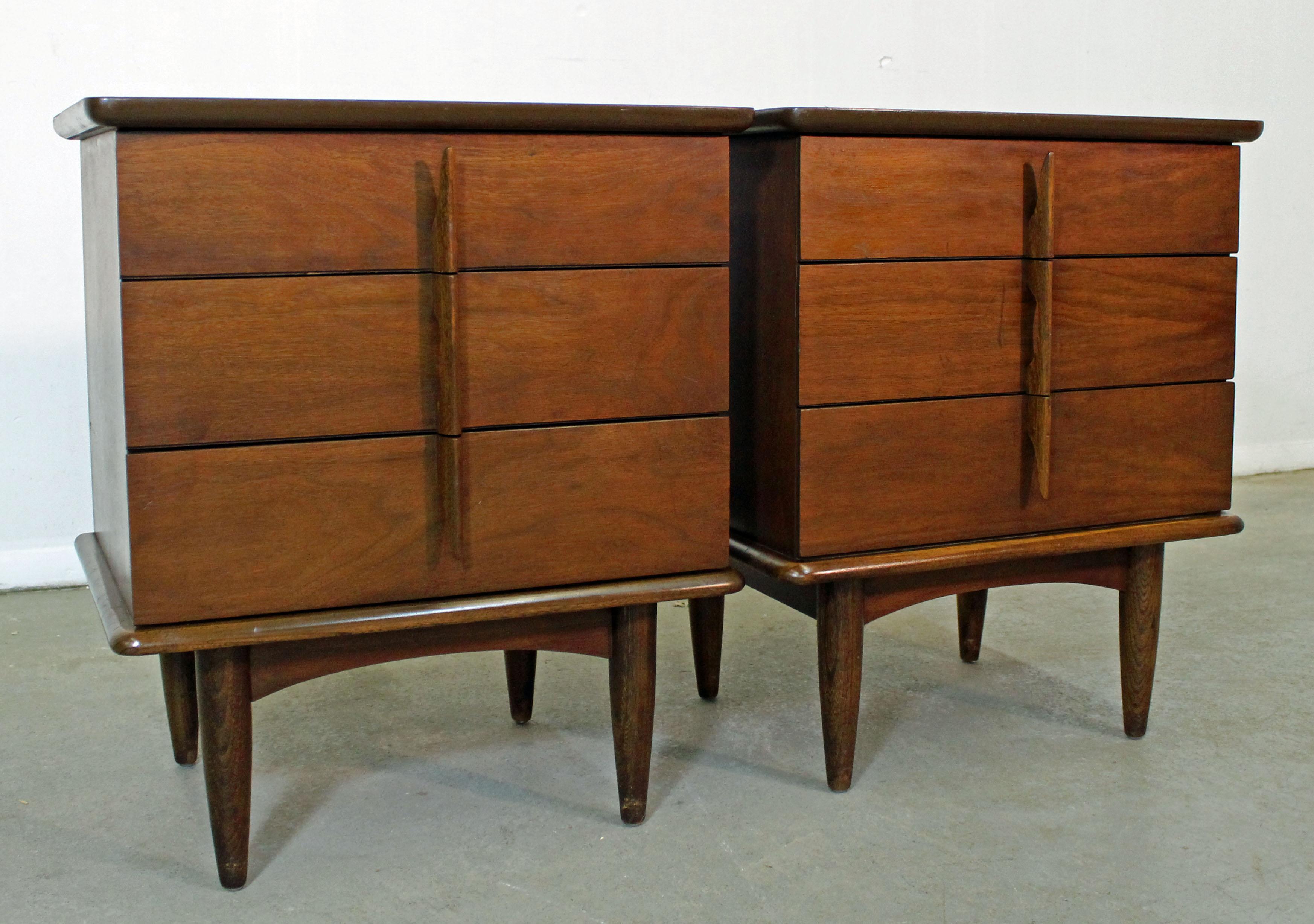 Offered is an excellent example of American Mid-Century Modern design. Includes a pair of walnut nightstands with three drawers and sculpted pulls. They are in good condition, tops have been refinished, but some show some age wear. They are not