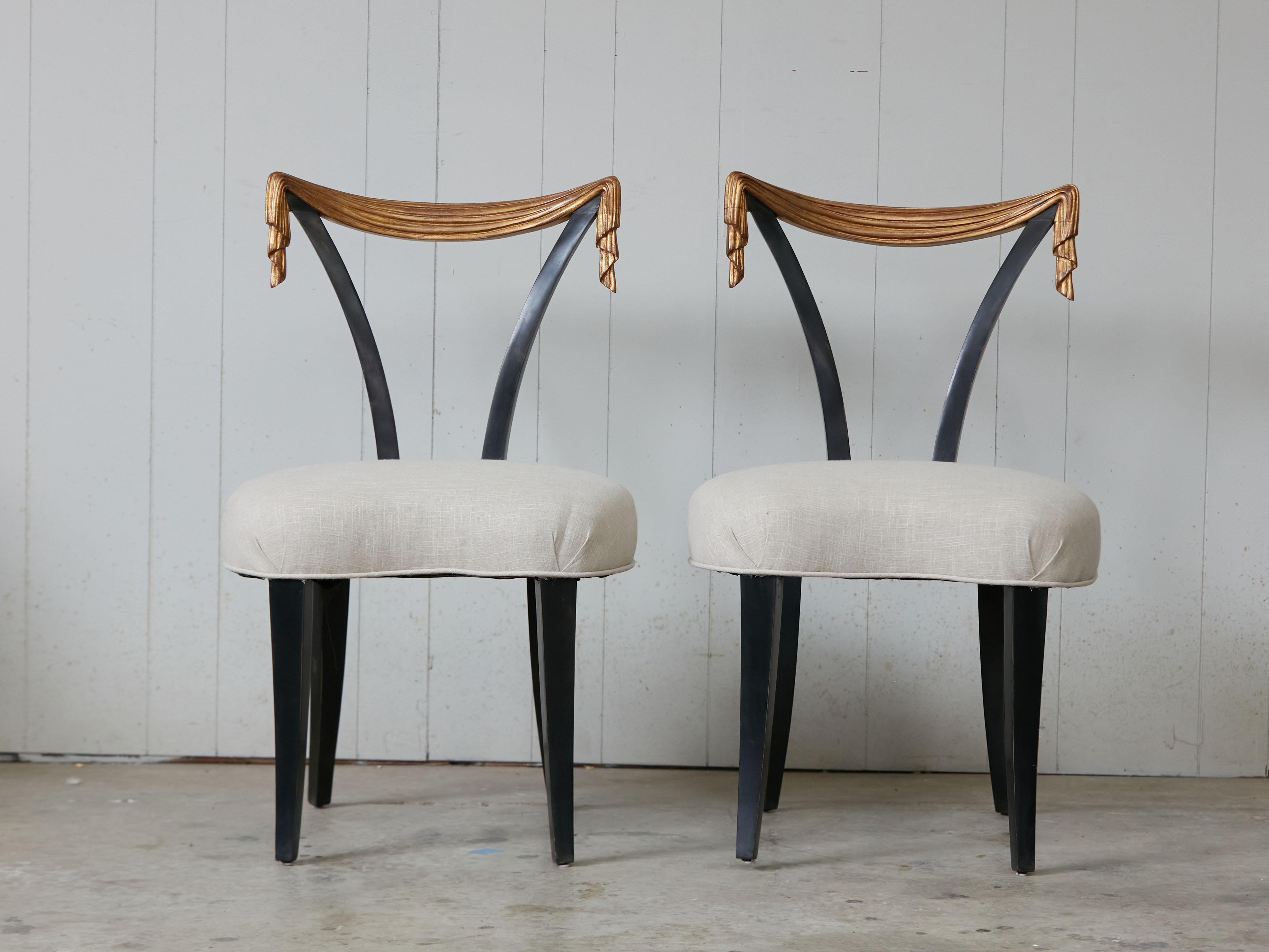 A pair of Dorothy Draper style Hollywood Regency black and gold side chairs from the mid 20th century, with hand-carved gilt draped backs. Charming us with their dramatic presence and contrasting colors, each of this pair of Dorothy Draper style