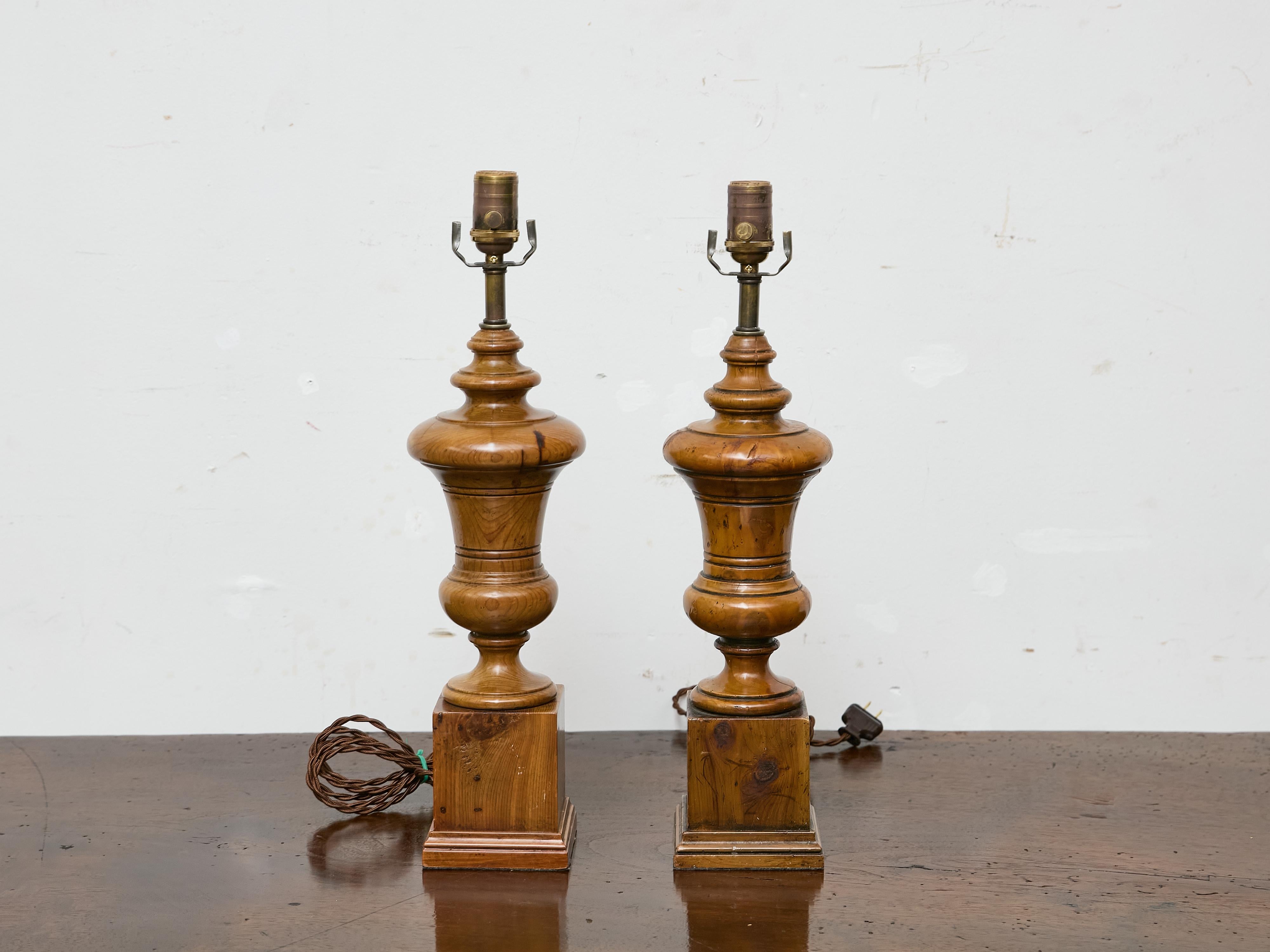A pair of turned pine table lamps from England, Midcentury, professionally rewired for the USA. Made of solid pine with a warm honey-toned finish and visible knots throughout, each lamp sits on a stepped square plinth that gives the base weight and