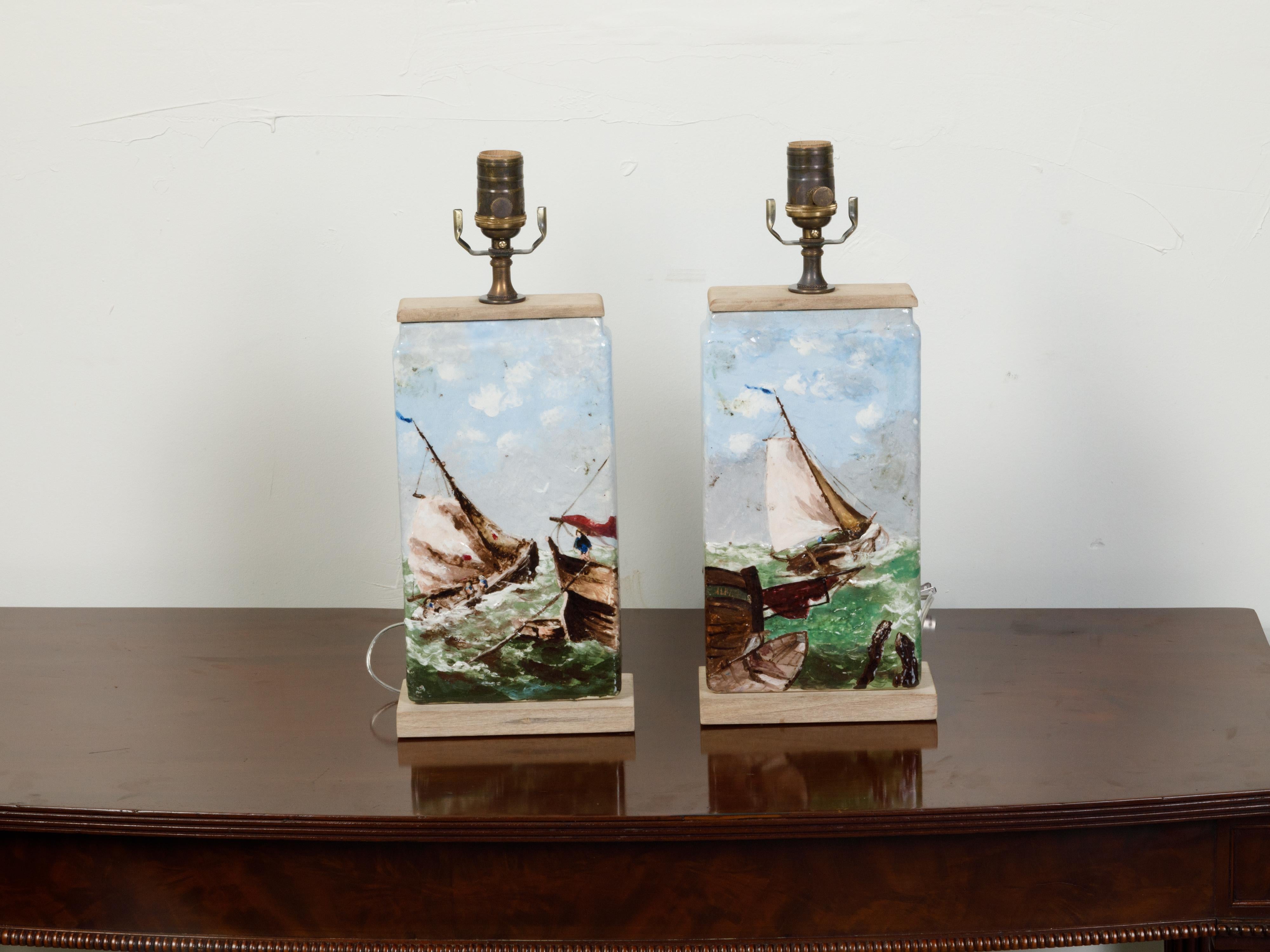 A pair of faience tables lamps from the mid 20th century, with hand-painted naval décor. Created during the midcentury period, each of this pair of table lamps features a rectangular faience body adorned with a hand-painted naval scene. Resting on