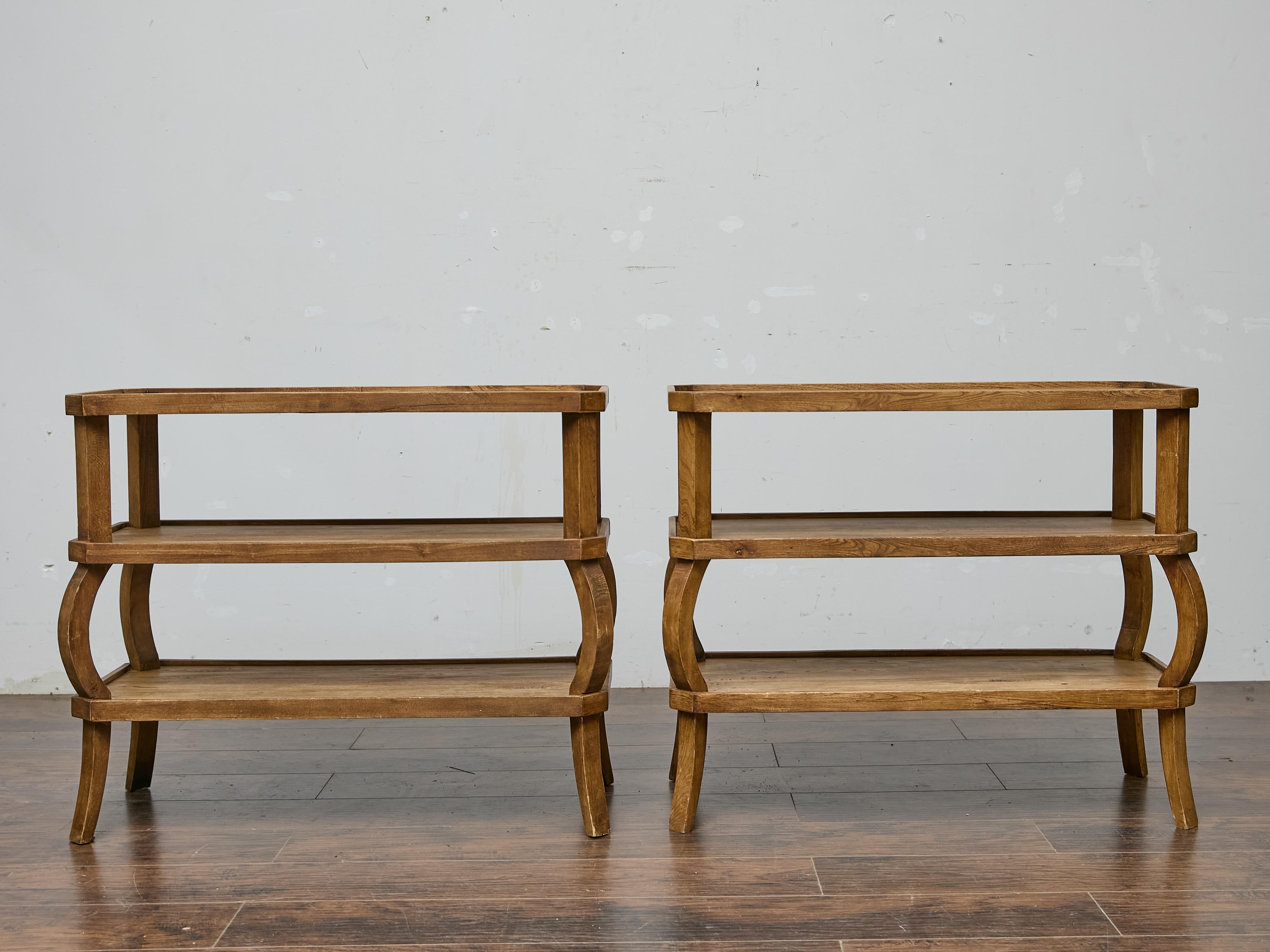Mit ihrer skulpturalen Silhouette und der warmen Patina bringt dieses Paar Stufentische aus französischer Eiche aus der Jahrhundertmitte Form und Funktion in Einklang. Mit ihren geschwungenen, architektonischen Beinen und den offenen Regalfächern