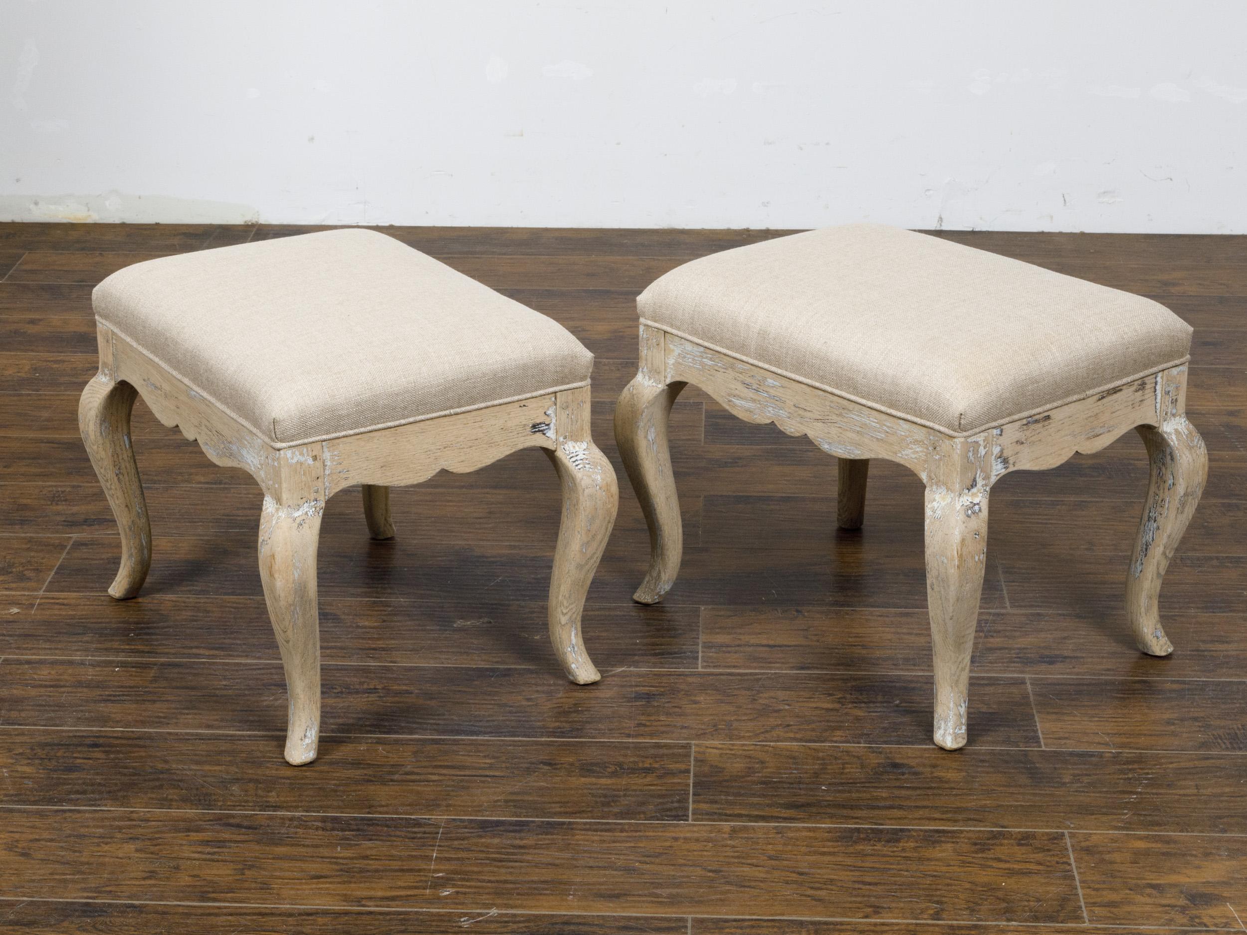 A pair of French Rococo style oak stools from the Midcentury period, with scraped back finish, scalloped aprons, cabriole legs and new custom linen upholstery. Imbued with the charm of the French Rococo style, this pair of mid-century oak stools