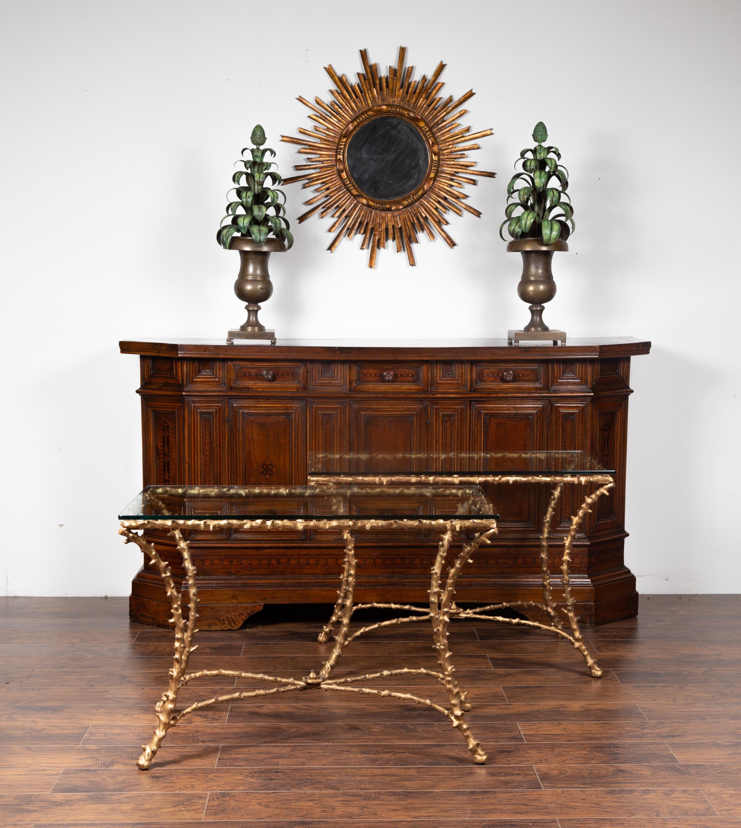 A pair of gilt bronze console tables from the mid-20th century, with faux bois motifs, glass tops and cross stretchers. Born during the midcentury period, each of these console tables attracts our attention with its stylish faux bois gilt bronze
