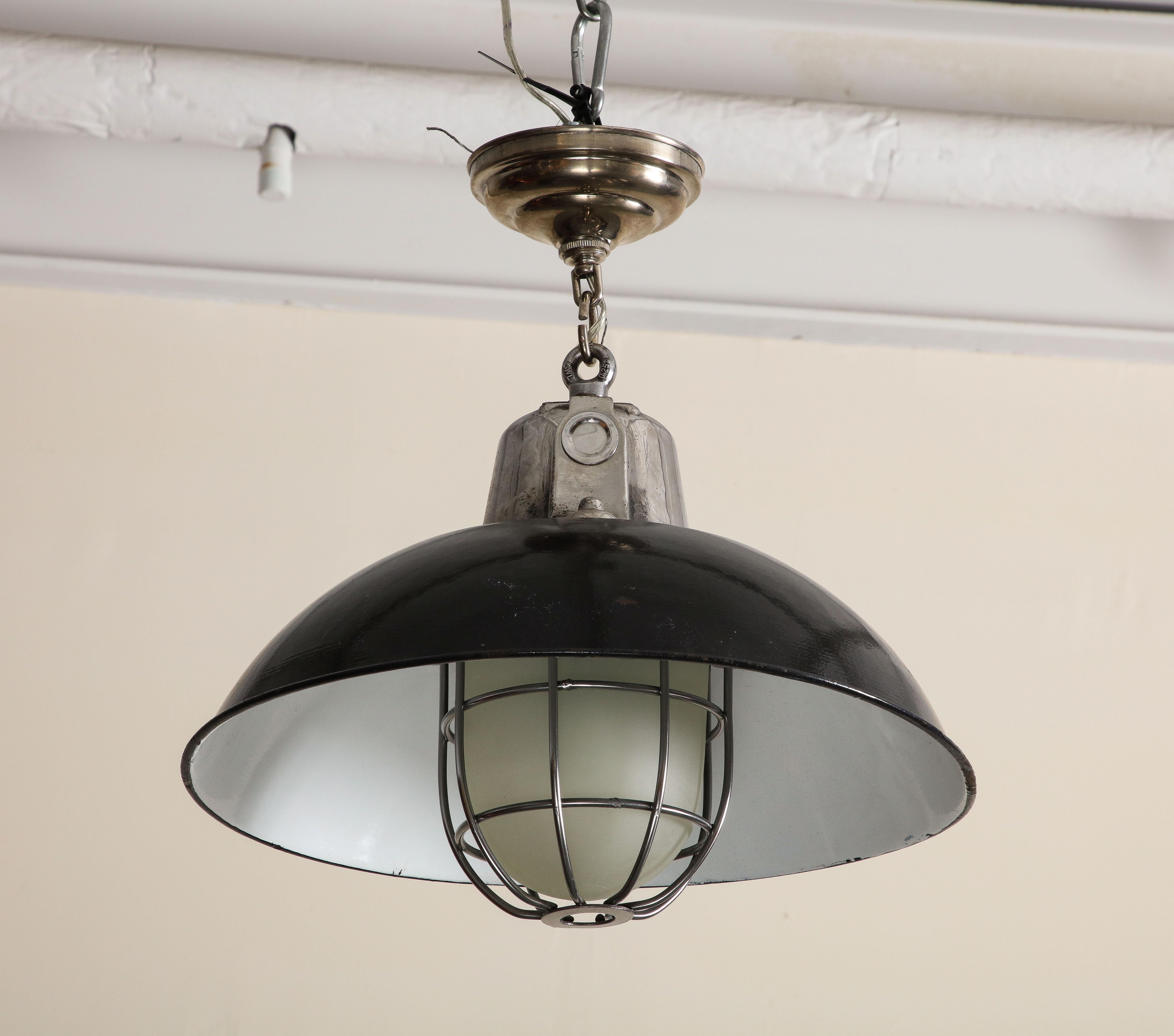 Pair of mid-century industrial style cage pendant lights with original black and white enamel. Wired for USA, good working condition. It features a black enamel shade, white enamel interior, cast iron top, frosted glass cover and iron grid.