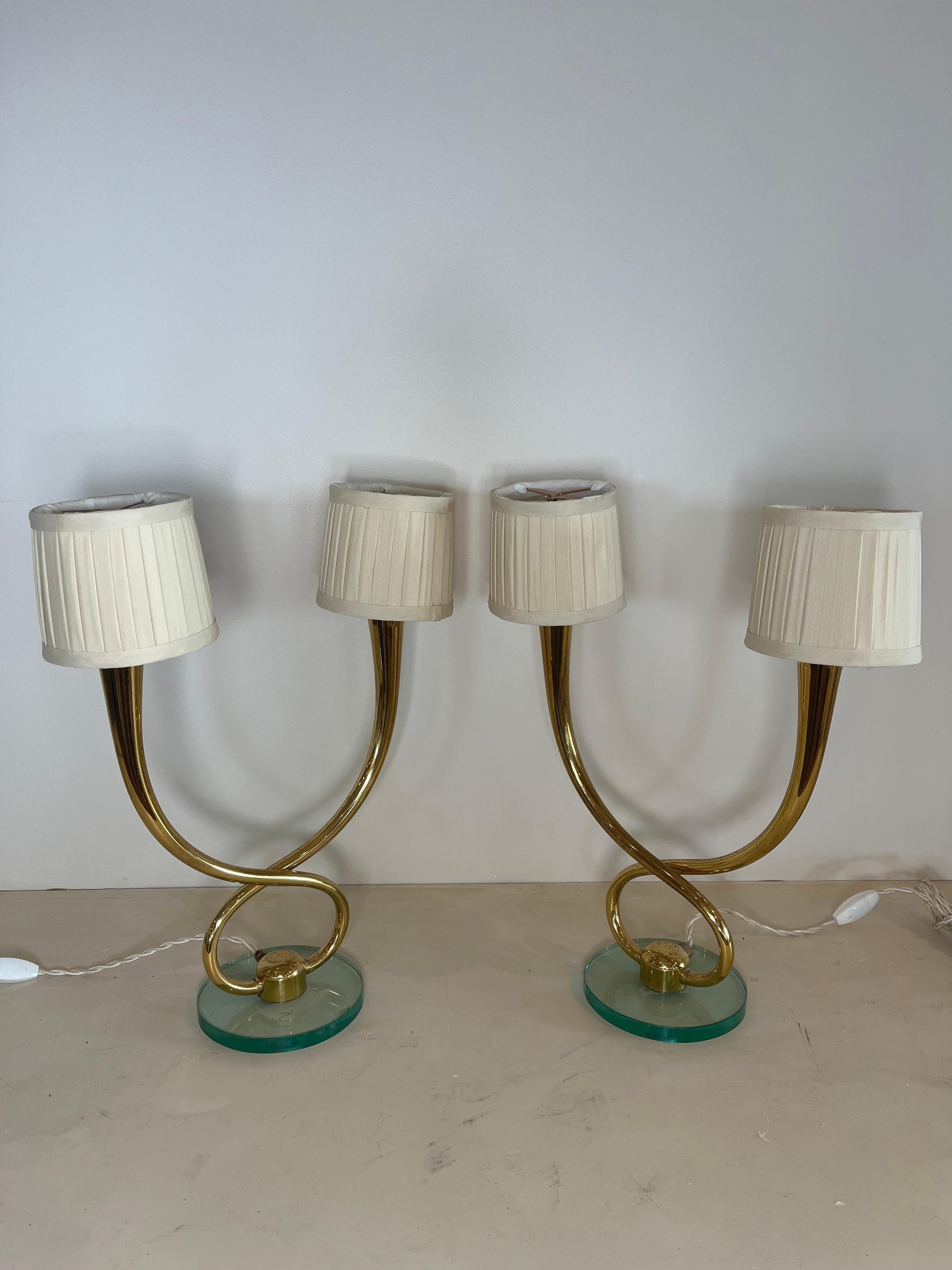 Pair of midcentury Italian table lamps, each with two lights on intertwined polished brass arms, stretching upward in a U-shape, and topped with ivory pleated shades. Base is a circle of molded glass. 

Newly rewired for USA, in good working