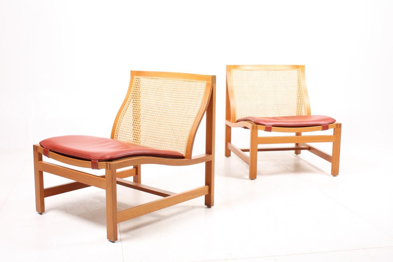 Pair of elegant lounge chairs with beech frame and French caning in seat and back. Seat cushion in patinated leather. Designed by Danish architects Johnny Sørensen and Rud Thygesen.