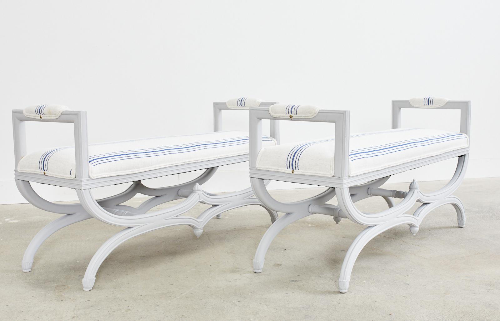 Matching pair of Mid-Century Modern lacquered window benches featuring curule style leg bases. The molded frames have a dove grey painted matte finish with newly upholstered seat cushions and padded arms. The soft fabric has a french linen blue and