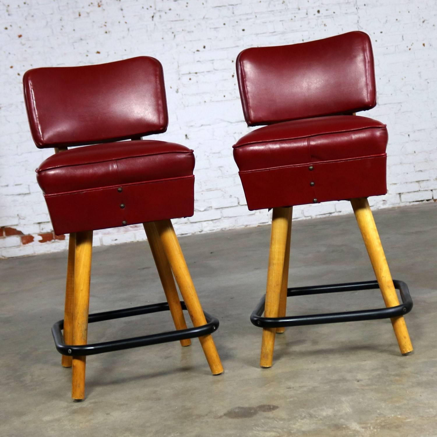 Awesome pair of midcentury counter height bistro bar stools in a deep read vinyl faux leather and blonde wood legs made by WCI aka West Coast Industries. These barstools are in excellent vintage condition apart from a three-corner tear in the