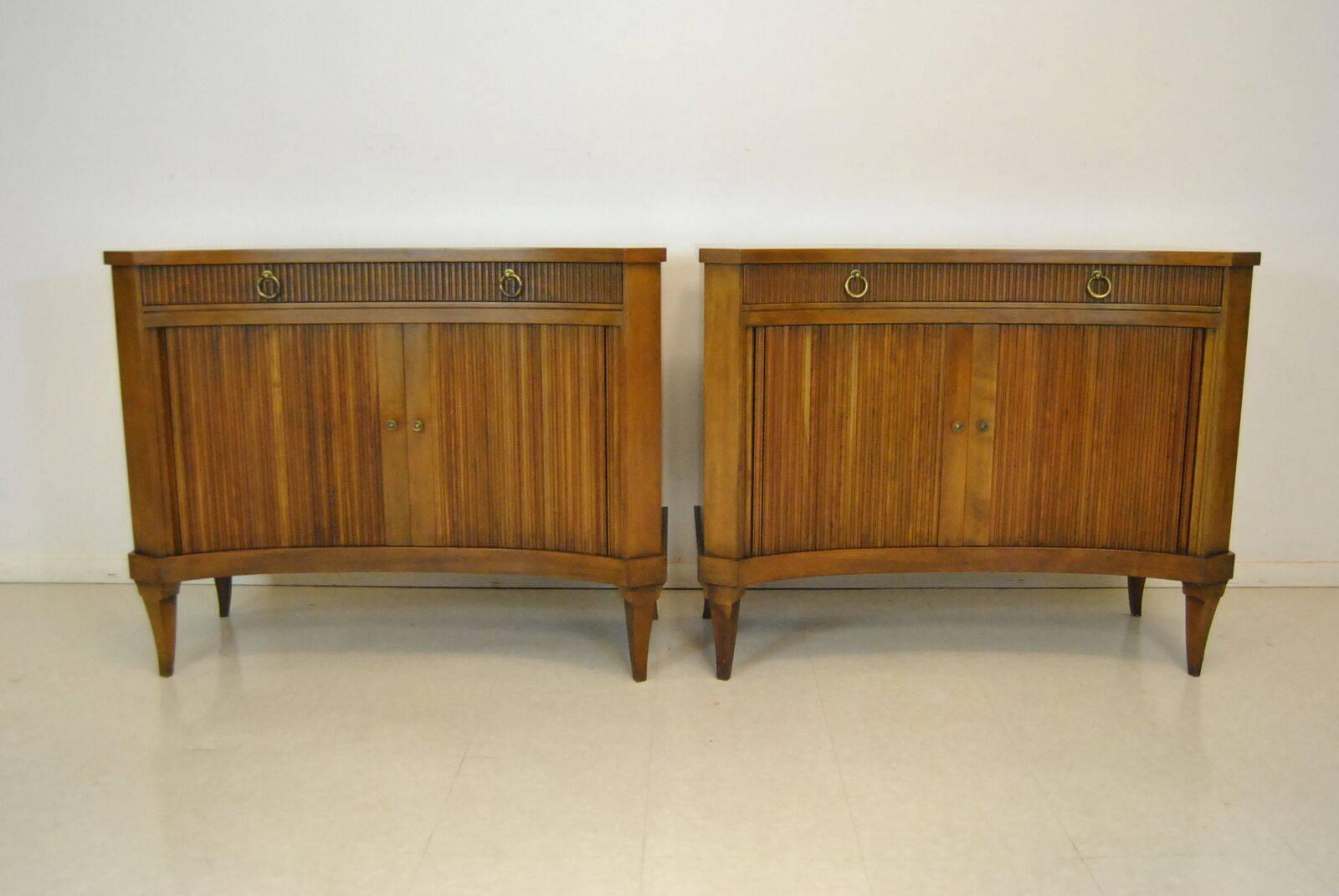 A very sought after pair of midcentury Regency style curved tambour door chests/commodes by Baker Furniture. These chests are made of solid fruit wood featuring a curved front with canted corners and tambour doors. They offer an interior bottom