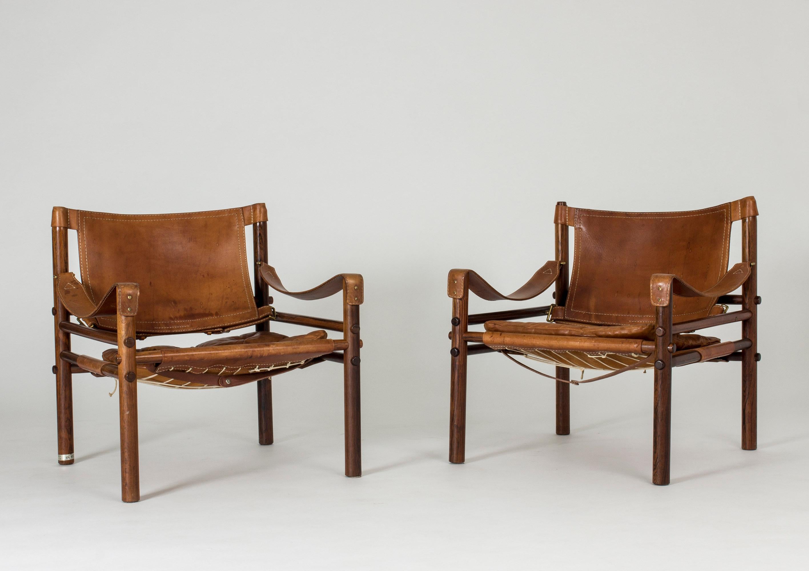 Pair of “Sirocco” lounge chairs by Arne Norell. Rosewood frame and cognac leather upholstery, with a removable cushion. Great details of wood plugs decorating the screw holes, seams in the leather and brass buckles. Very nice patina.