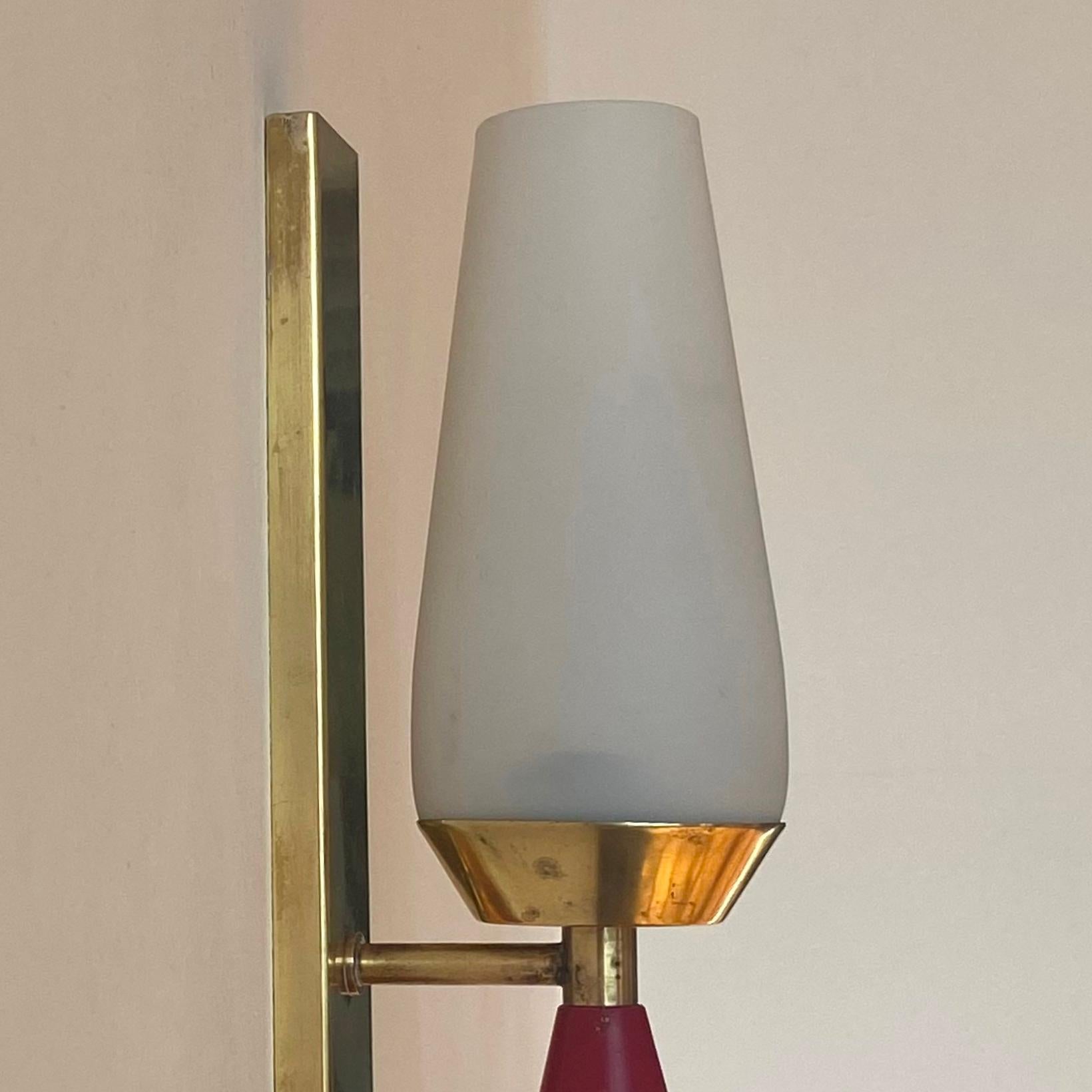 Con reminiscencias a la manera de Stilnovo, el aplique se fabricó  en italia en  la década de 1960. Los materiales utilizados en la fabricación del candelabro son latón, laca roja y cristal opalino.  La lámpara está en condiciones de funcionar.