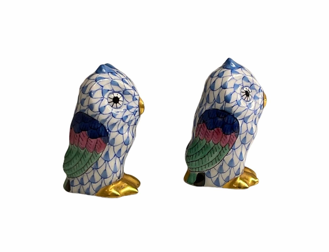 This a pair of Herend porcelain hand painted miniatures owls figurines. They are painted white in the background with royal blue fish scale pattern. They have large black eyes, gilded peaks and feet. Their feathers are painted blue, pink and green.