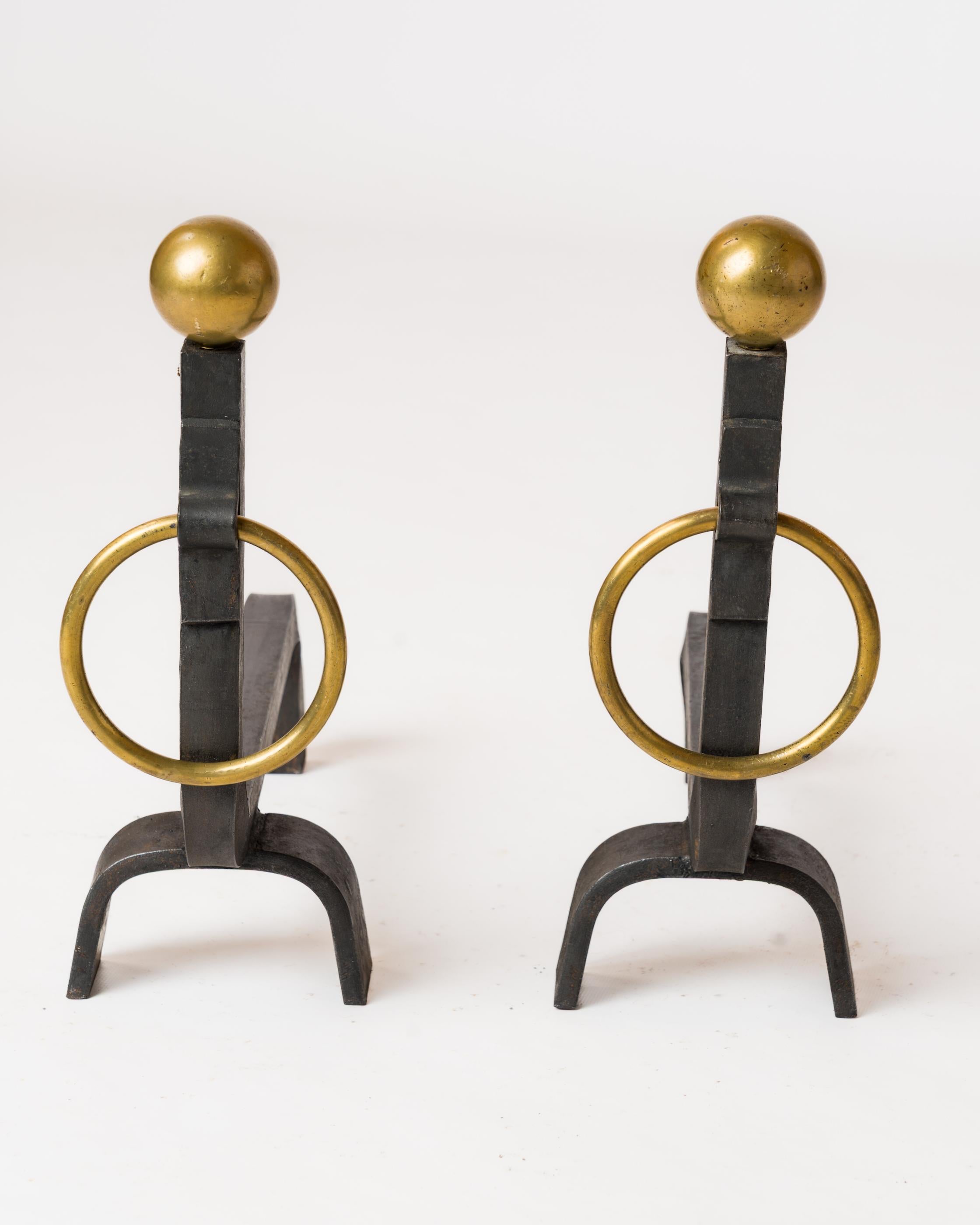 Chic and minimalist pair of wrought iron andirons with polished brass hoop handles and finishing balls. In fair vintage condition. 
These andirons will ship from France and can be returned to either France or to an upstate NY facility. 
Price