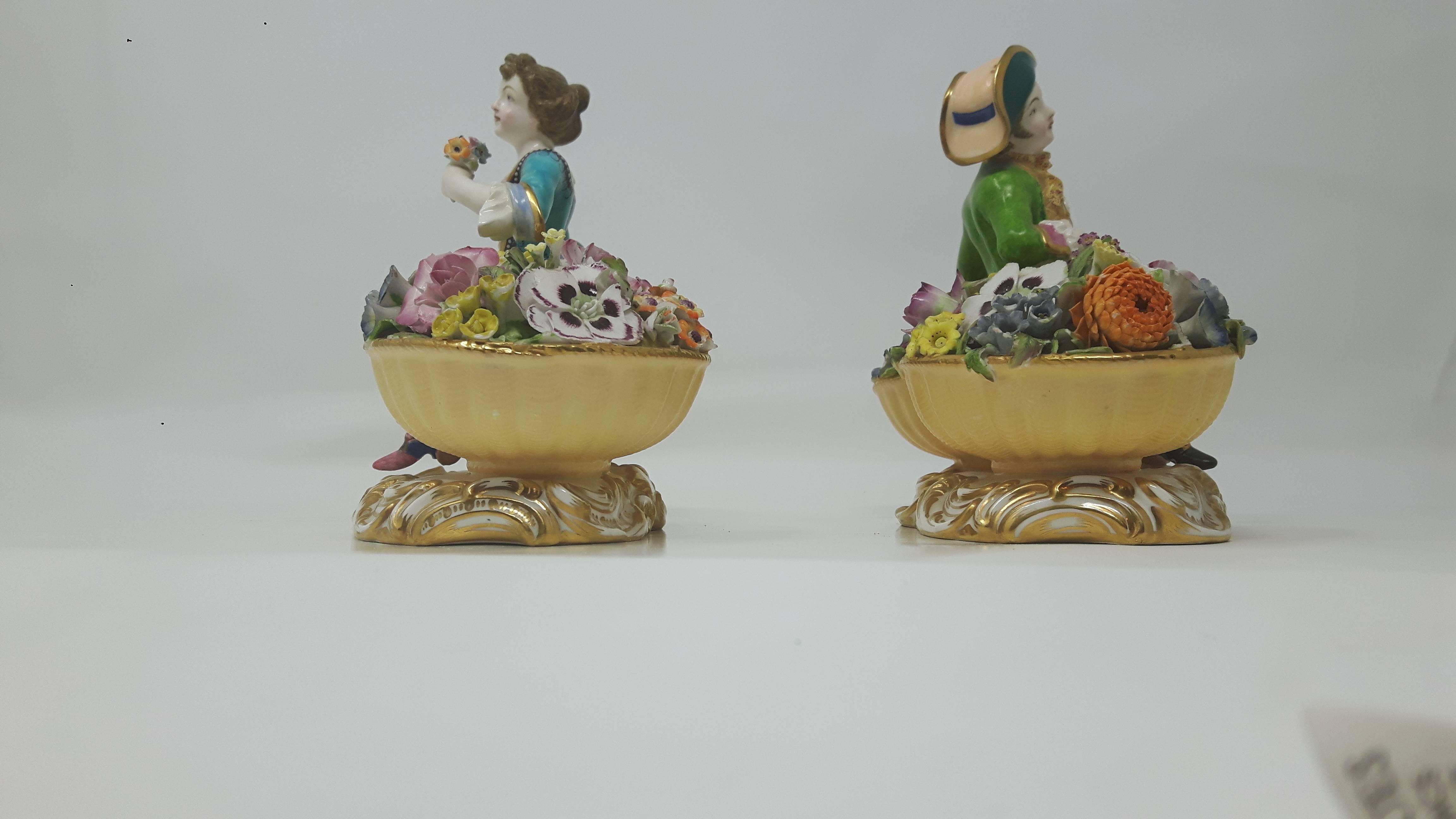 A pair of Minton figures of flowers sellers in the form of a youth and a girl in 18 century costume, each seated between two large baskets of very fine flowers, on gilt scrolled bases, model numbers 58 and 59 ,circa 1830-1840.