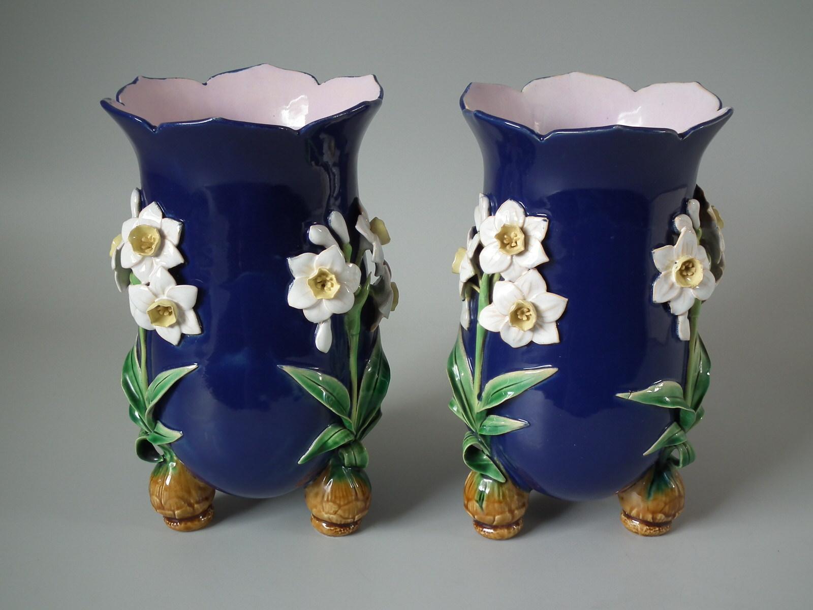Pair of Minton Majolica vases which feature daffodils stood on bulb feet. Cobalt blue ground version. Colouration: cobalt blue, green, white, are predominant. The piece bears maker's marks for the Minton pottery. Bears a pattern number, '1200'.