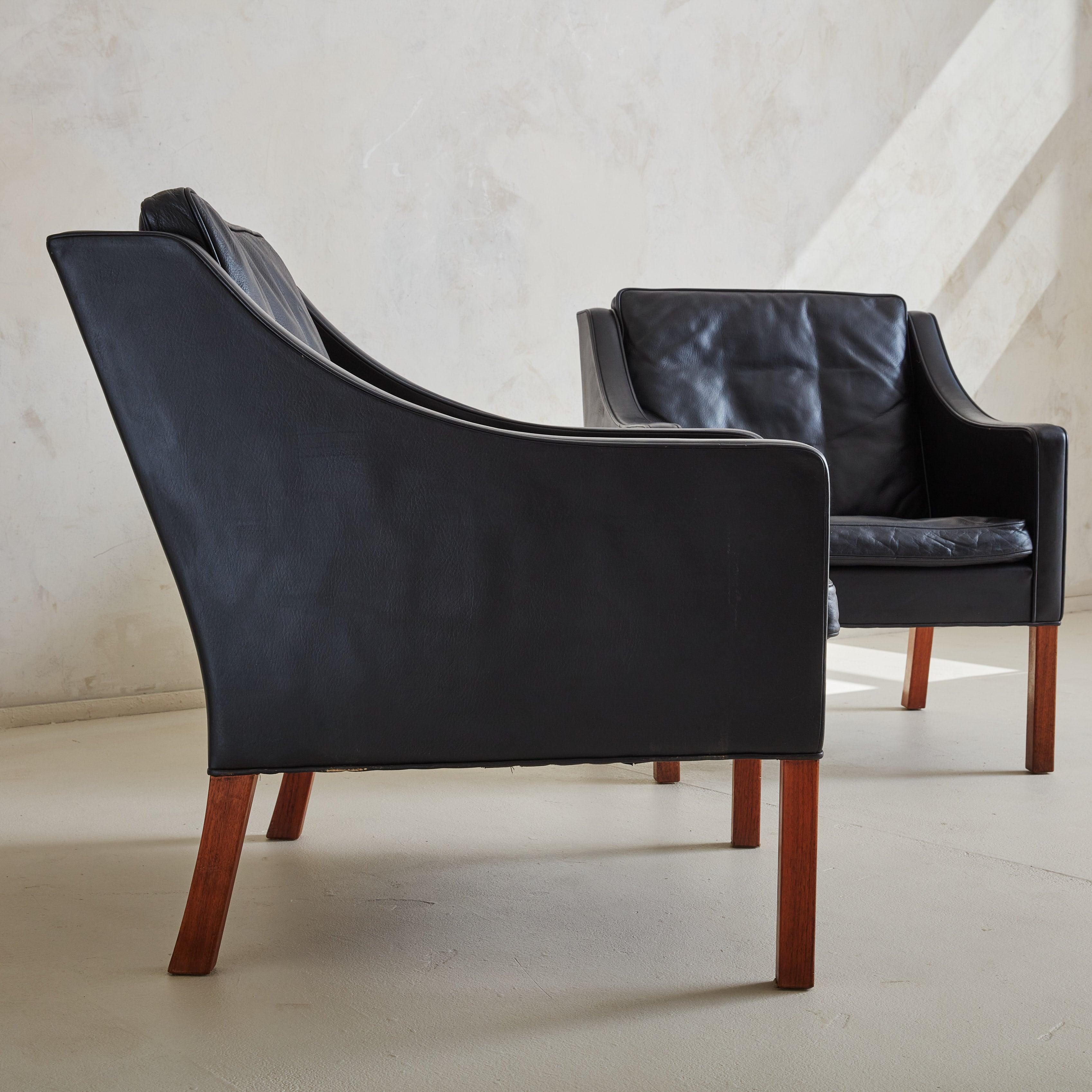 A pair of Mid-Century Modern ‘Model #2207’ lounge chairs upholstered in black leather with two removable down-filled cushions. The structured silhouettes feature elegantly sloped arms, curved backs, and minimalistic, solid wood legs in a warm,