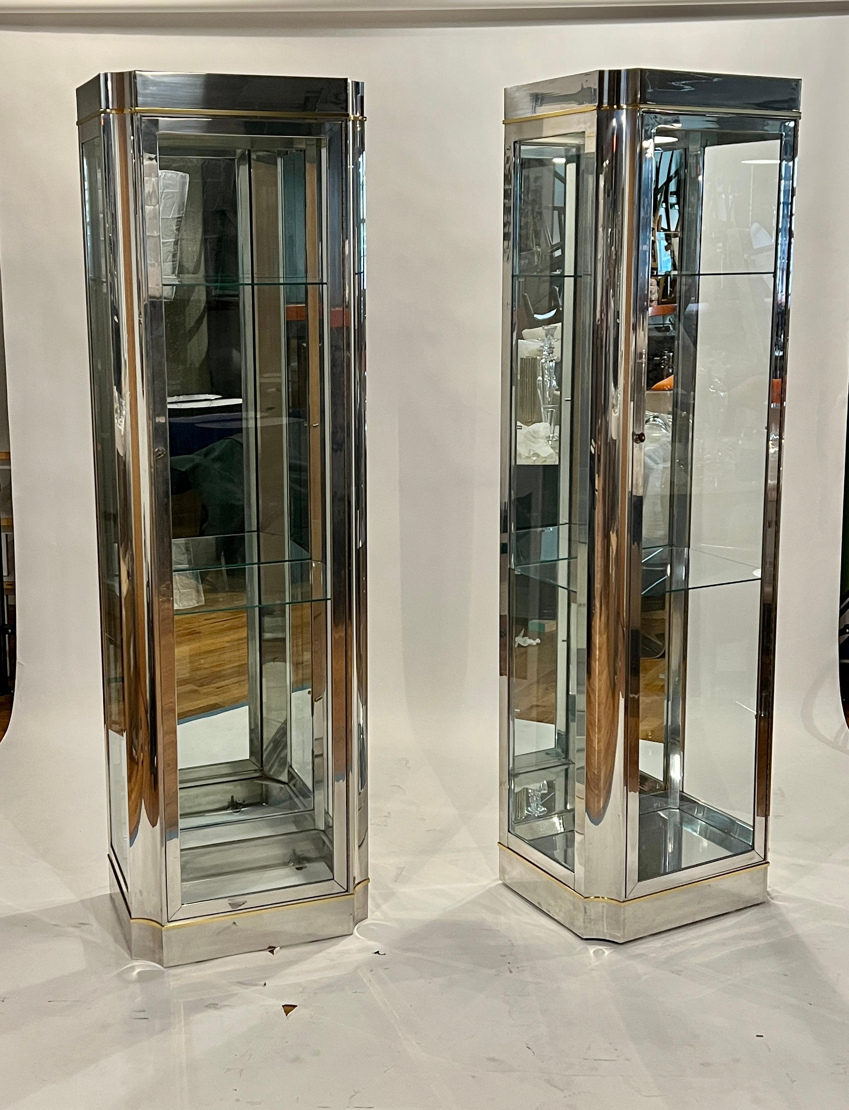 Pair of 20th century vitrines after Mastercraft made in chrome and glass with brass accents. Each cabinet features one door which opens to 5 glass shelves which may be removed if desired. The cabinets illuminated from above to showcase your