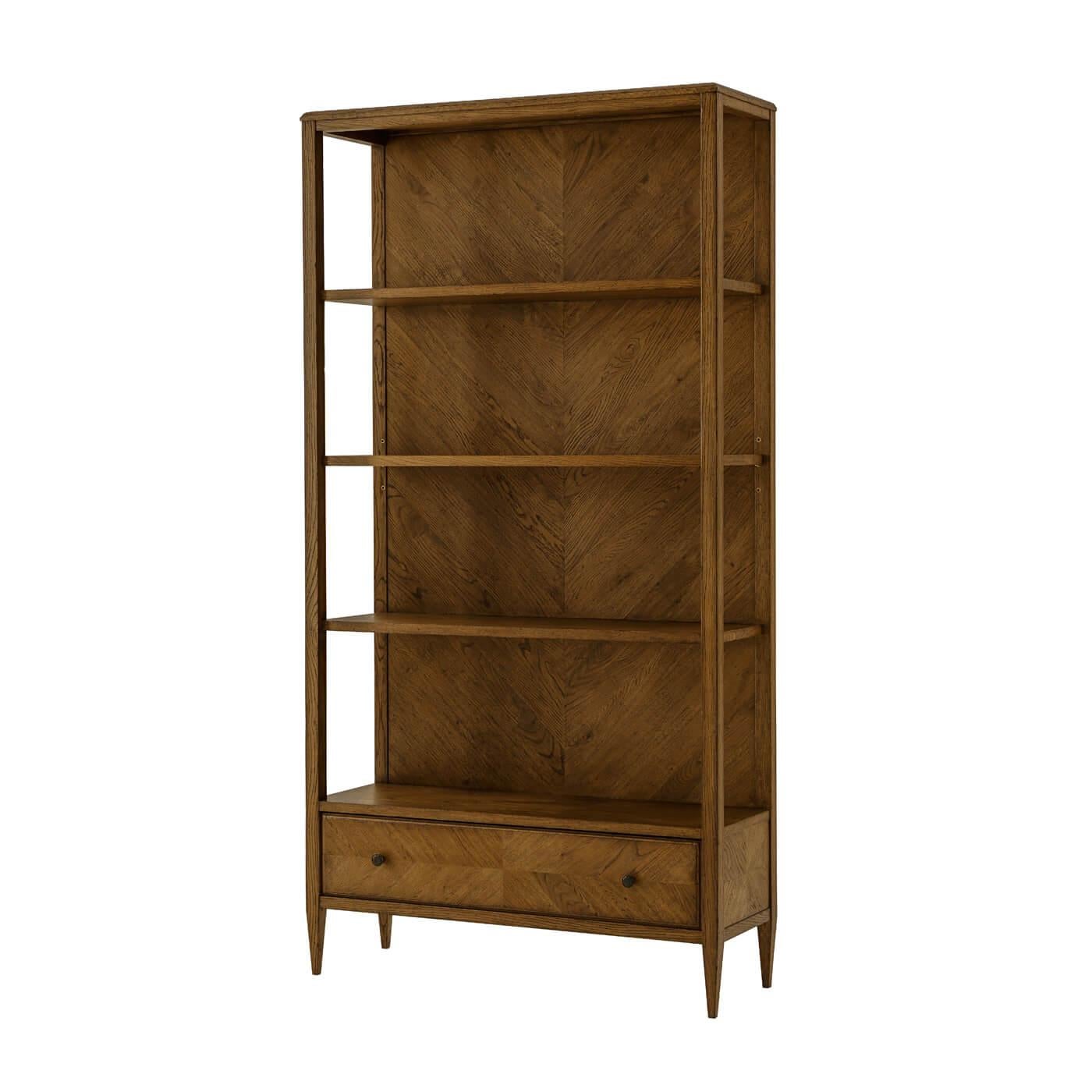 A Modern herringbone parquetry bookcase in our dark oak Dusk finish with three veneered shelves and a herringbone parquetry design on the bottom drawer. It sits on tapered legs with Verde Bronze finished knobs. A beautiful functional piece for
