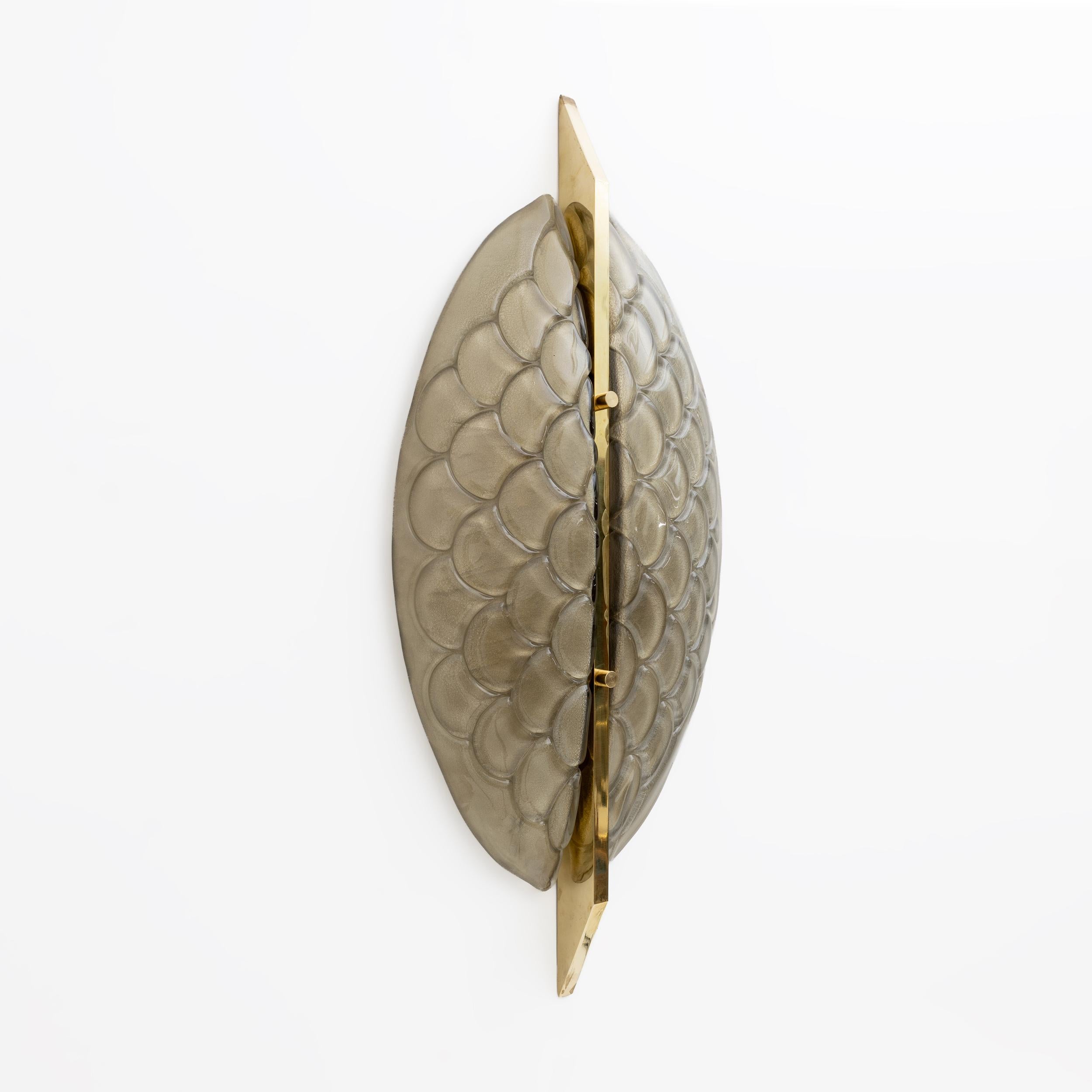 This pair of wall lamps features two thick glass structures: blown and pressed glass to obtain a fish scale effect, translucent and structured Murano glass, the glass extends vertically along the central brass motif. Overall, the lamps have an