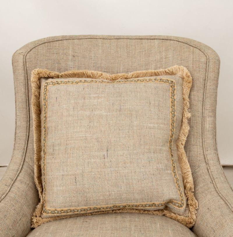 Paire de fauteuils modernes recouverts de tissu gris, avec coussins à franges assortis, apparemment non marqués. Le tissu est un tissage croisé dans des tons neutres de beige, de marron clair et de touches de gris. De subtiles taches bleues sont