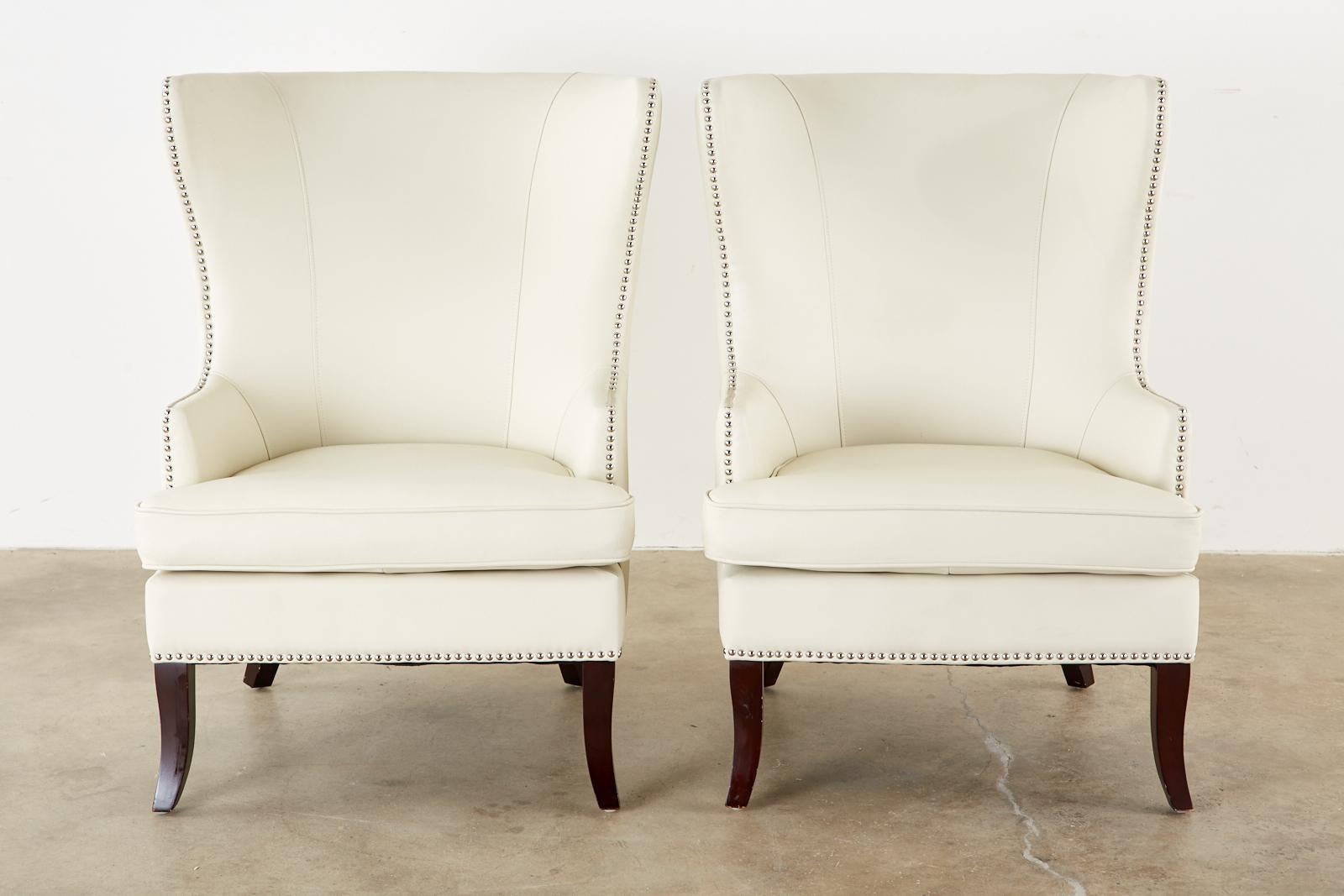 Stunning pair of modern style wing chairs or lounge chairs featuring a dramatic white leatherette upholstery. Crafted with hardwood frames and decorated with nickel finished brass tack nail heads on the borders. Fitted with a conforming loose seat