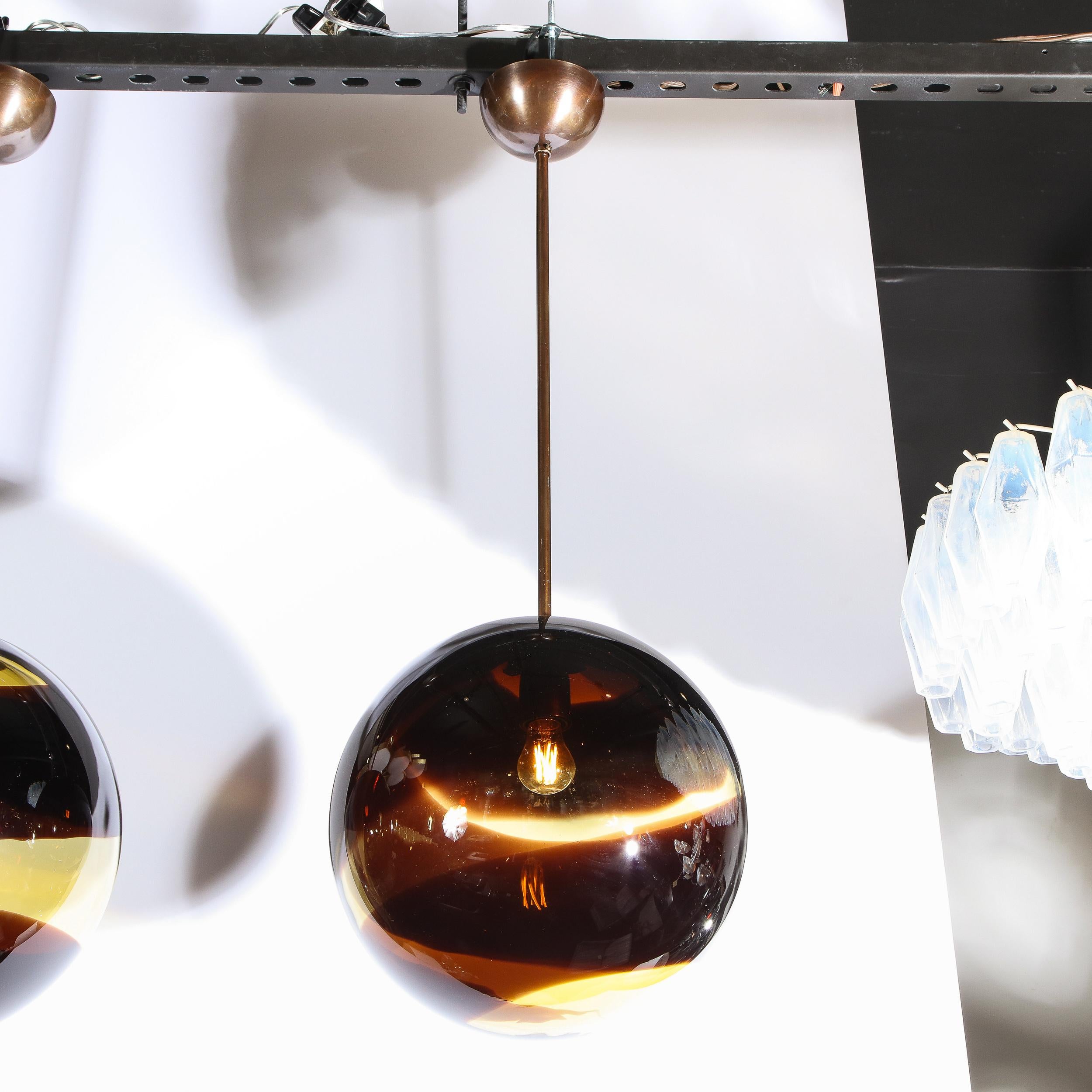 This elegant pair of modernist pendants were handblown in Murano, Italy- the island off the coast of Venice renowned for centuries for its superlative glass production. The fixtures offer spherical bodies in smoked amber Murano glass with undulating