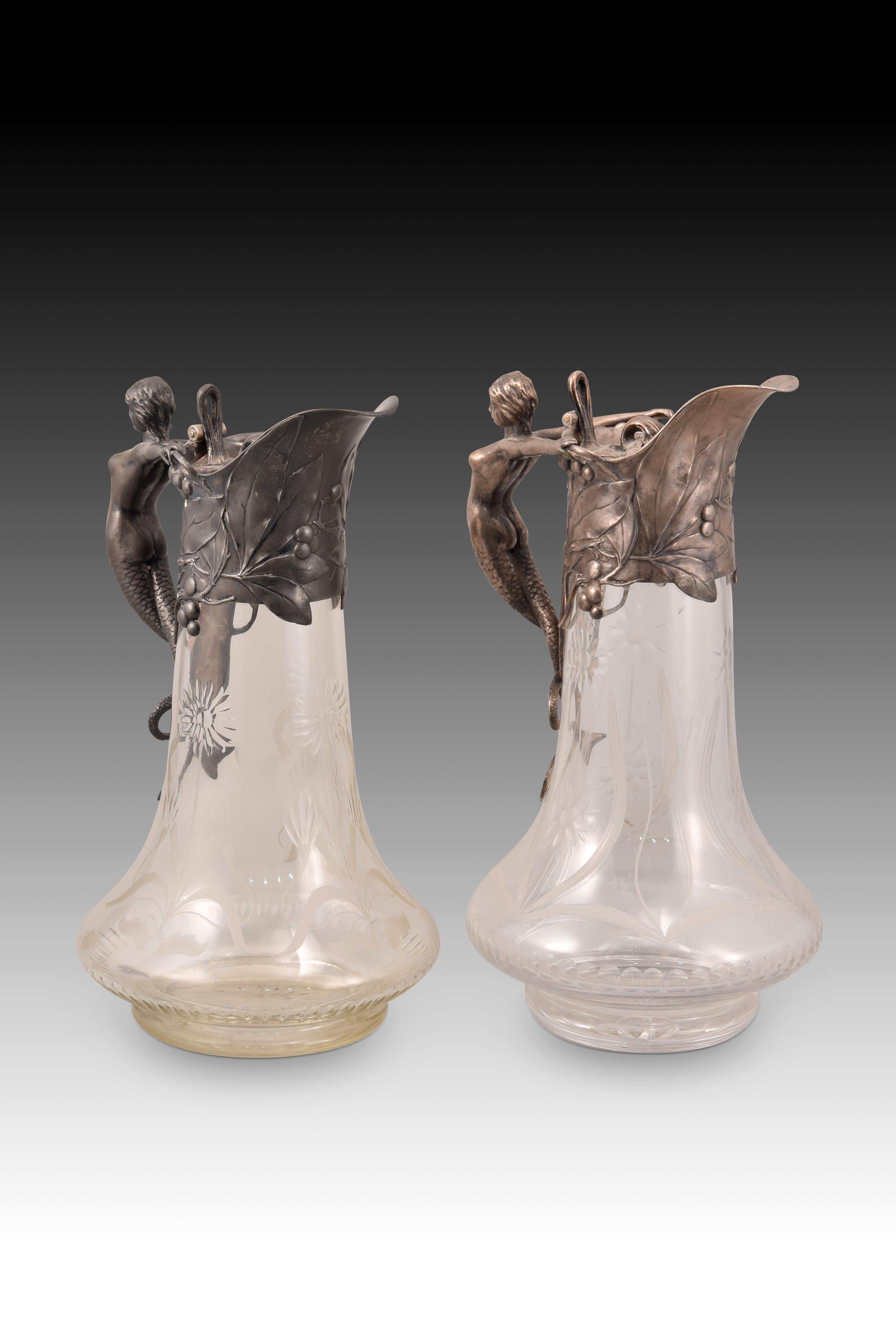 Pair of modernist jugs. Pewter, cut glass. WMF, Germany, 1903. 
They show marks. 
Pair of jugs with transparent etched and cut glass body and handle and top with silver pewter lid. The pewter area presents a delicate work of leaves, stems and