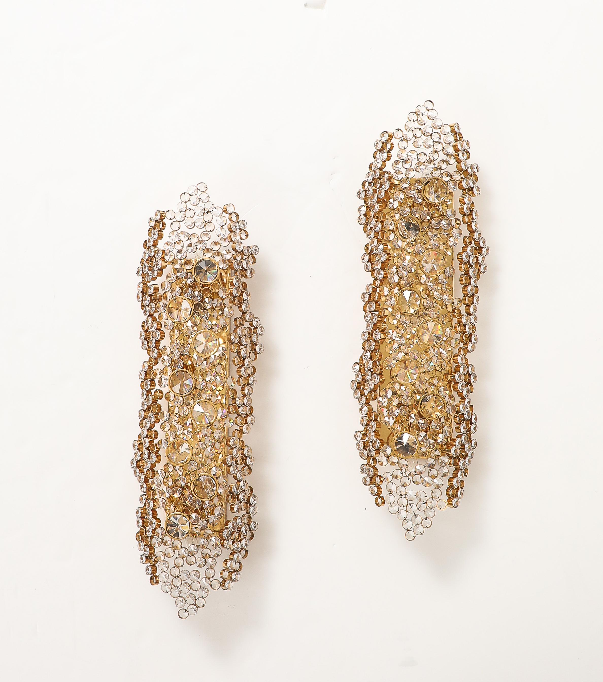 Espectacular pareja de monumentales apliques de latón dorado y cristal de la década de 1960, obra de Palwa, de la serie Luz Encantada.
Cientos de anillos de latón dorado con incrustaciones de cristales de distintos tamaños forman juntos un panel