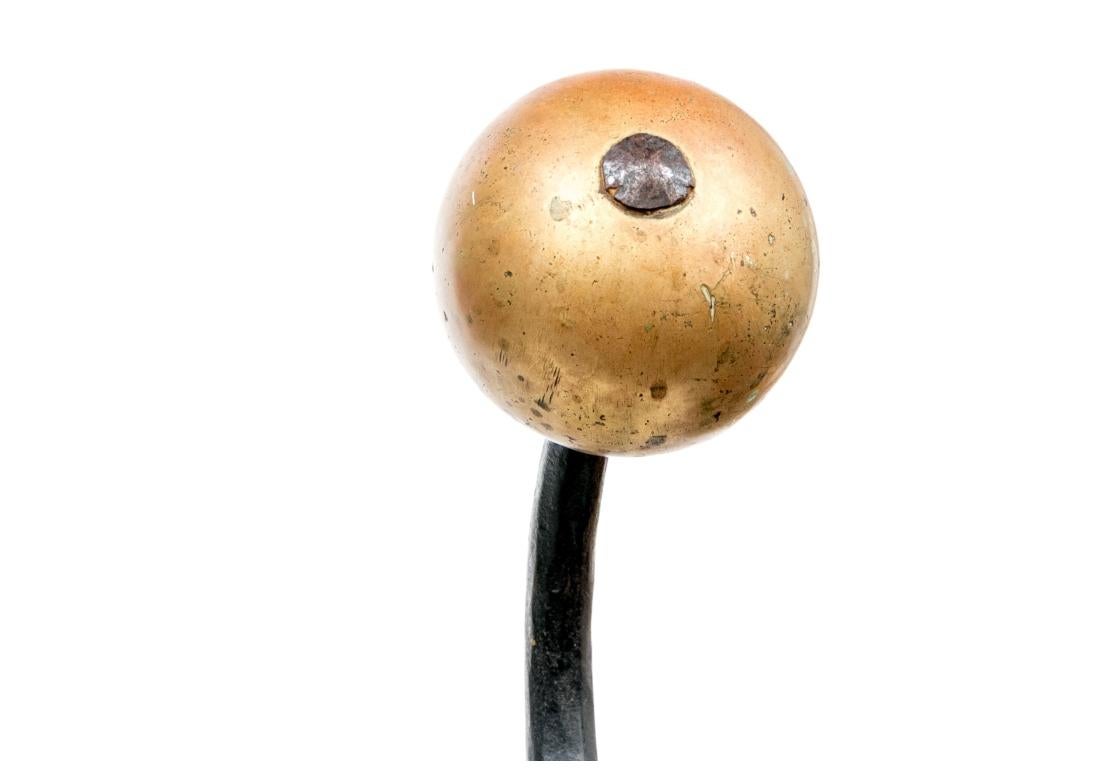 Wrought iron andirons in the Spanish Colonial Revival style. These monumental andirons are designed to hold logs and pots within a fireplace and feature decorative elements including large brass globe finials, brass rings, swinging bracket and