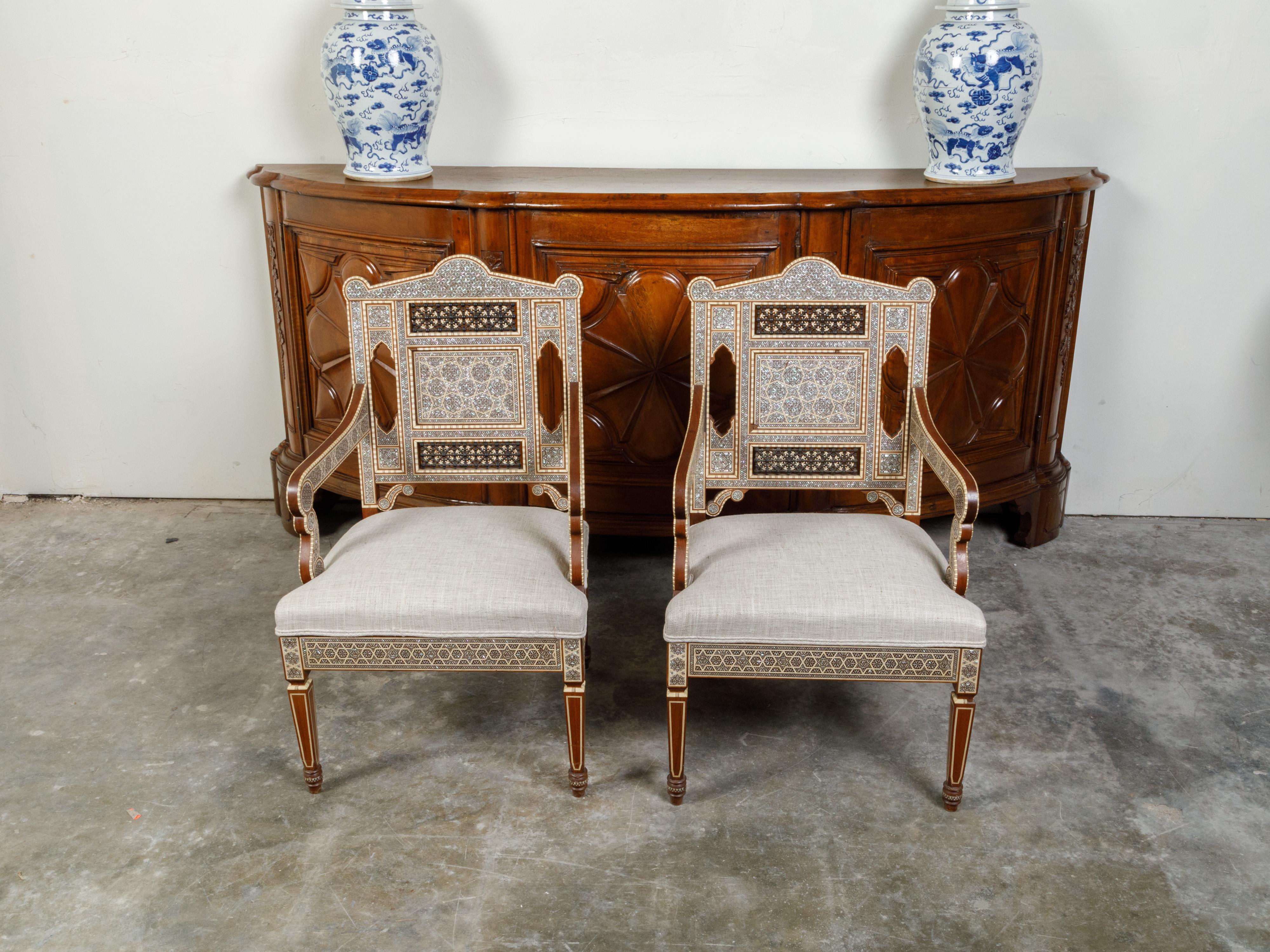 A pair of Moroccan armchairs from the mid 20th century, with geometric mother-of-pearl inlay and new upholstery. Created in Morocco during the mid-century period, each of this pair of armchairs captures our attention with its stunning back adorned