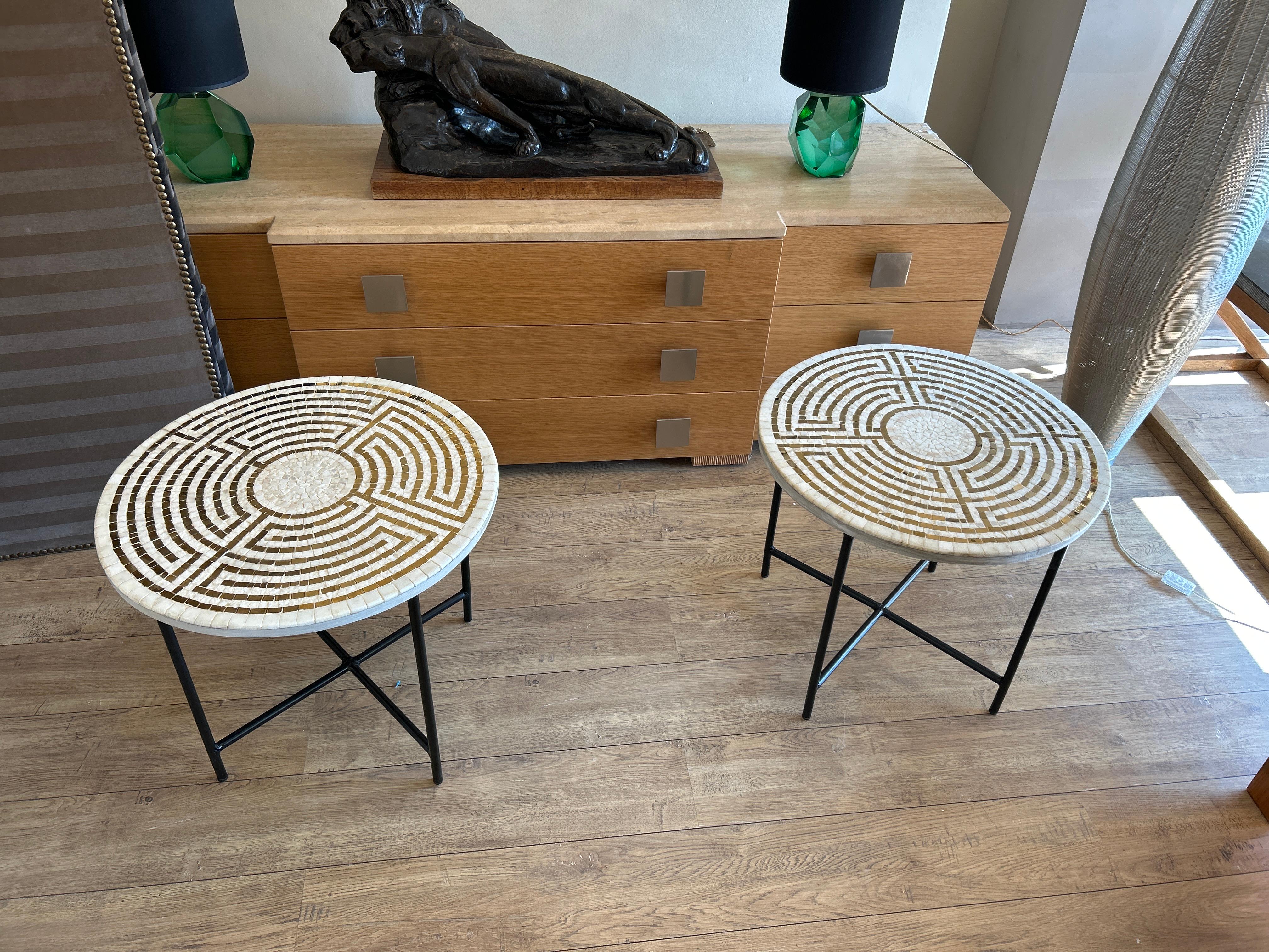 Pareja de mesas auxiliares de mosaico. El motivo se compone de teselas blancas y doradas dispuestas en círculos concéntricos para formar un laberinto geométrico, creando un efecto moderno y artesanal a la vez. Su base de metal negro añade un toque